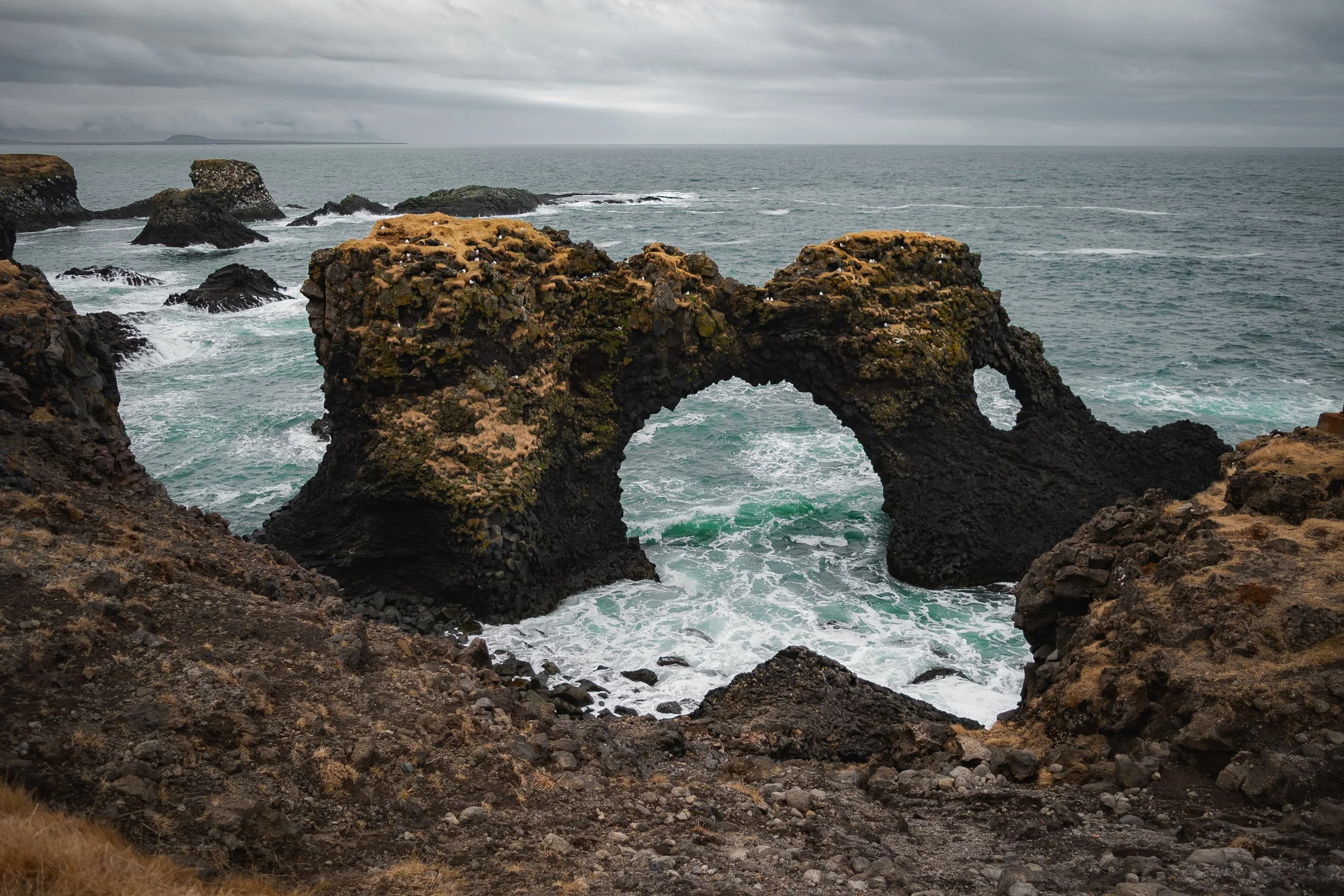 Coastal scene with rocky cliffs and natural arch over the ocean, under cloudy sky.