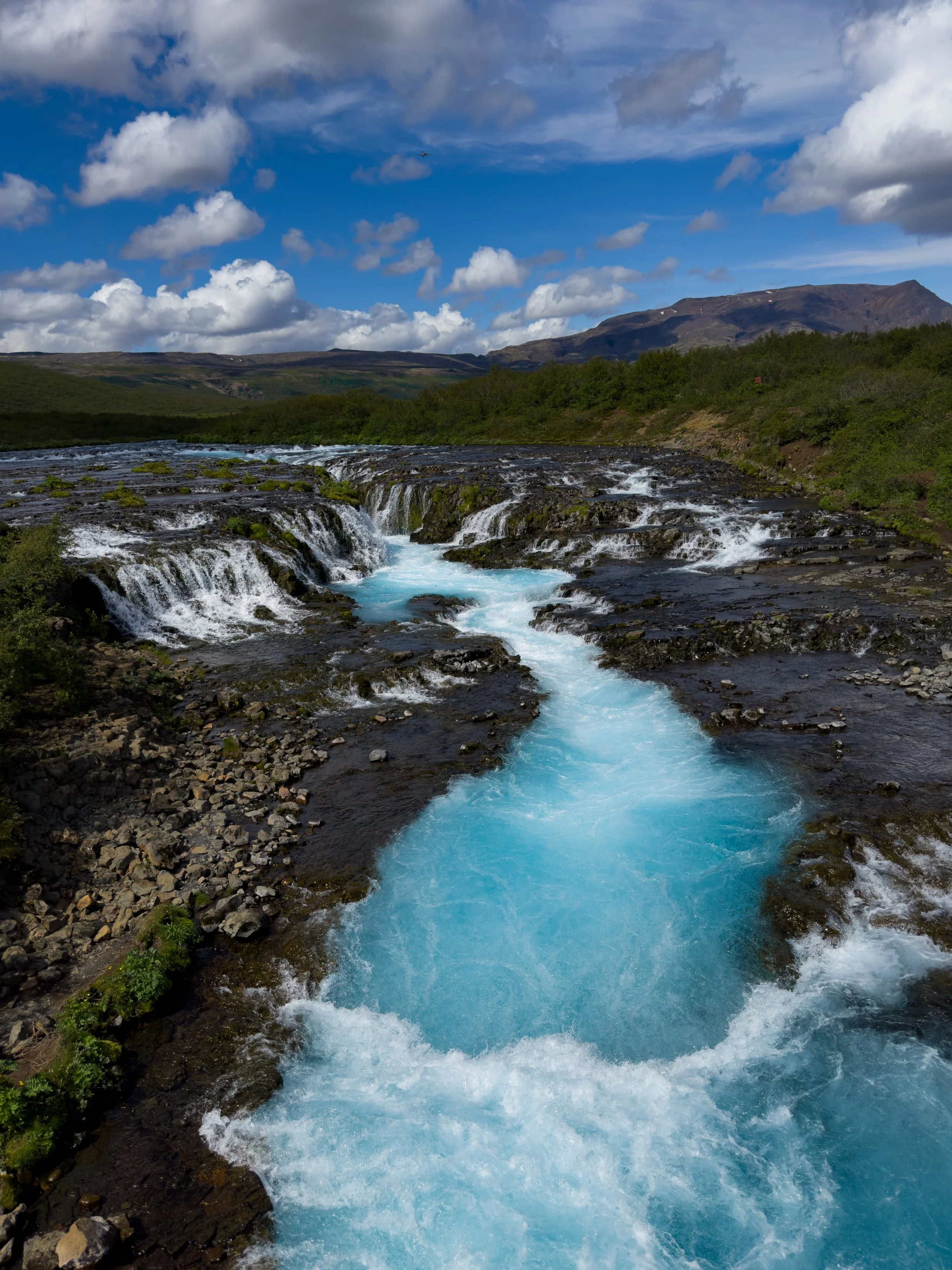 A river with turquoise water flowing over rocks, surrounded by green bushes under a sky with scattered clouds and mountains in the distance.