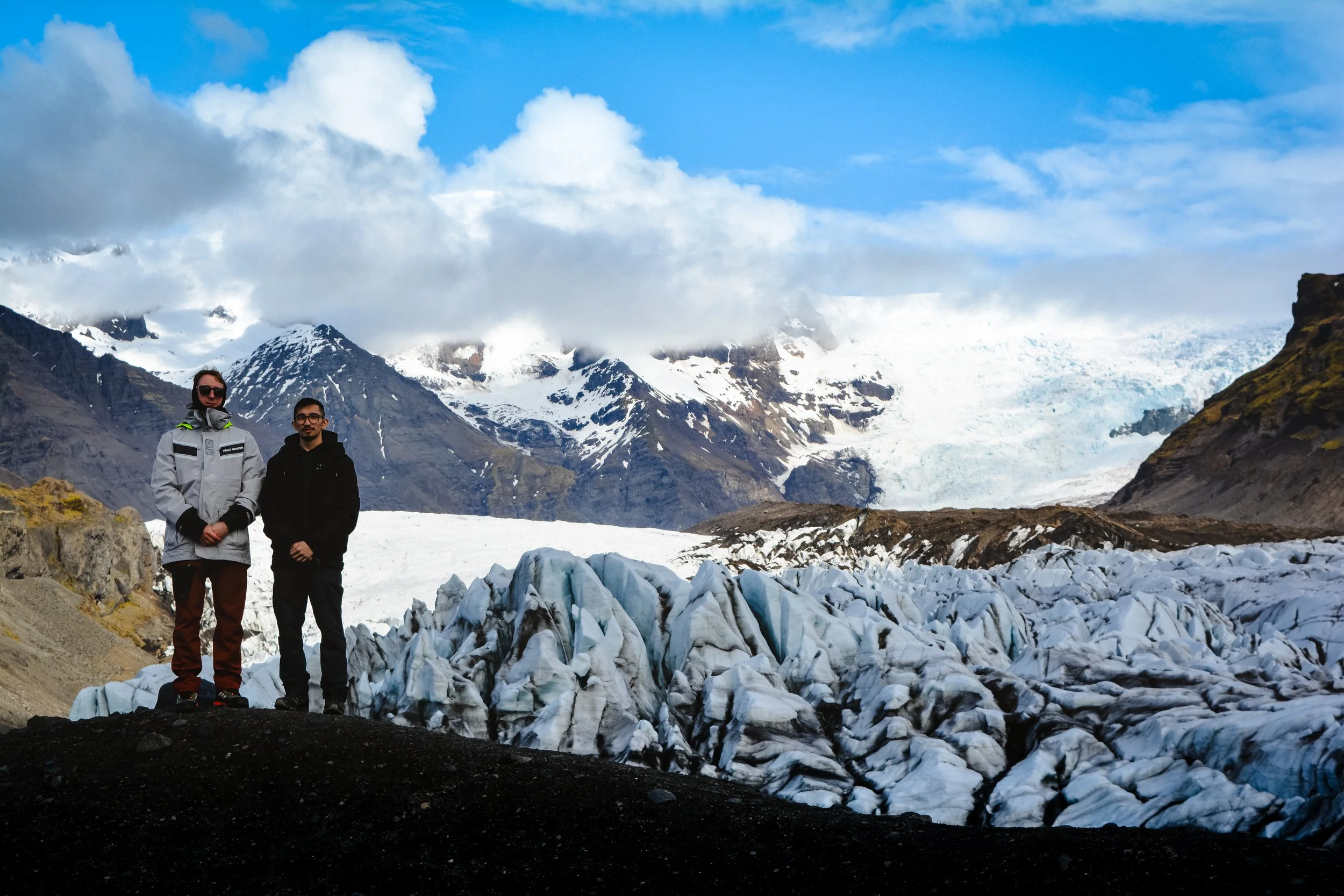 Two people standing on rocky terrain surrounded by snow and glacial ice with snow-capped mountains and a partly cloudy sky in the background.