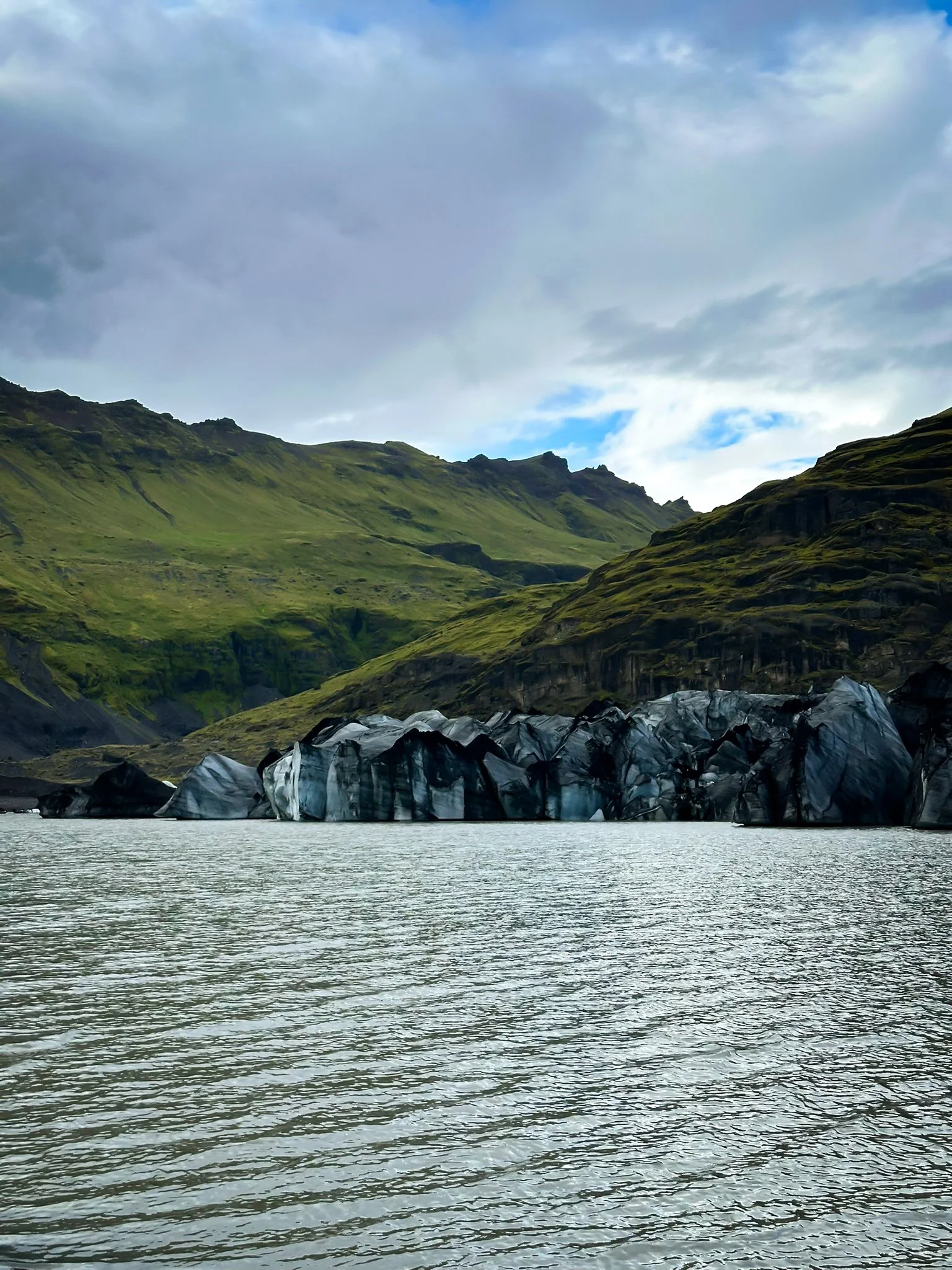 A landscape with a body of water in the foreground, rocky outcroppings, and green mountains with some rugged peaks under a partly cloudy sky.