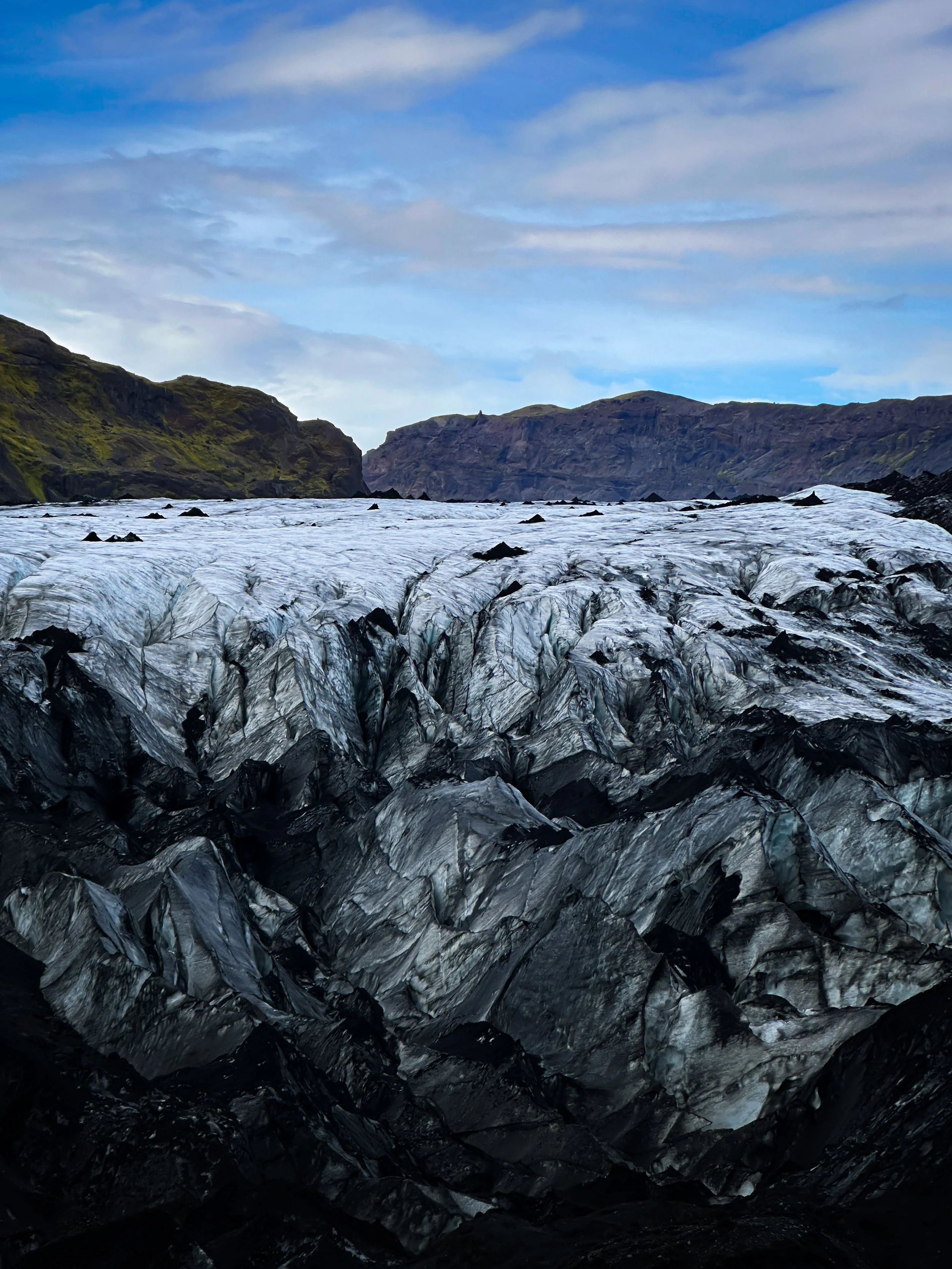 A glacier with black volcanic rocks at the front, surrounded by green hills, under a partly cloudy sky.