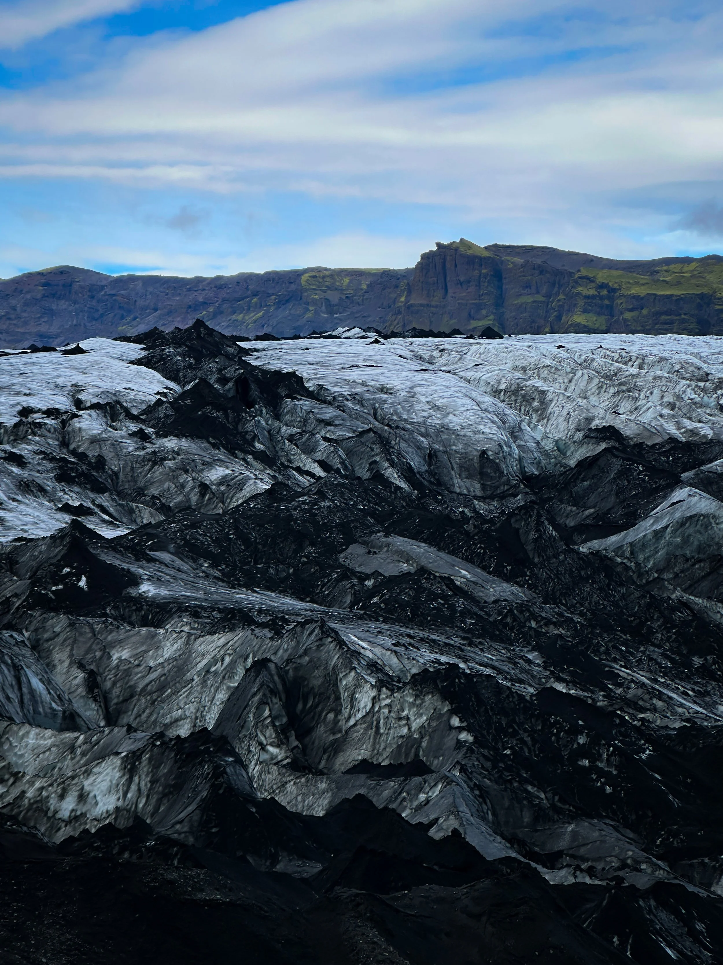 A glacier with black volcanic rocks on its surface, set against mountainous terrain with green vegetation and a cloudy sky.