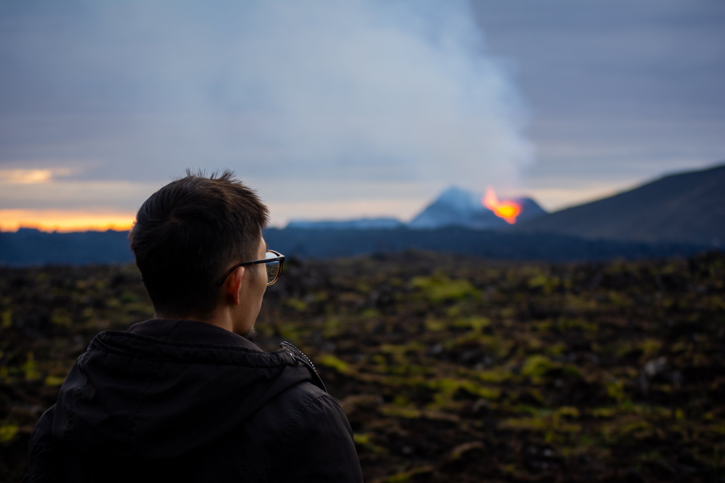 Man with glasses looking at erupting volcano with lava and smoke, during dusk.