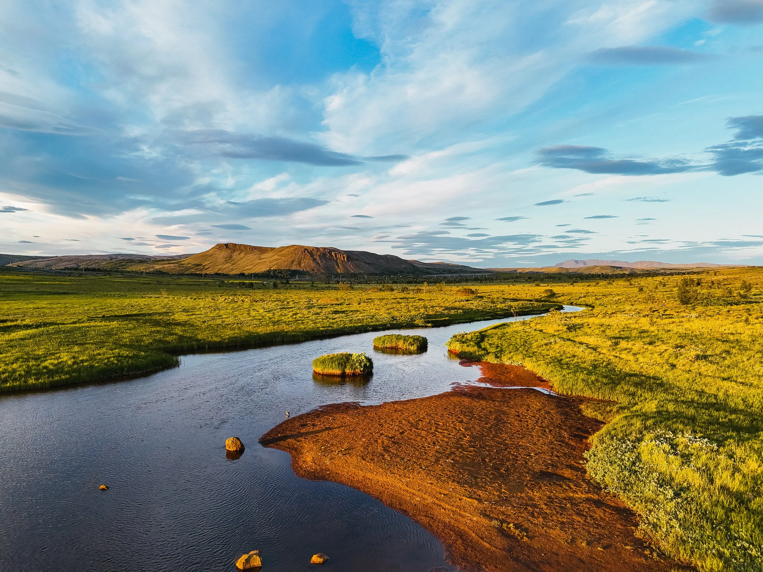 Sunset over a meandering river in a grassy plain with a mountain in the distance and a partly cloudy sky.