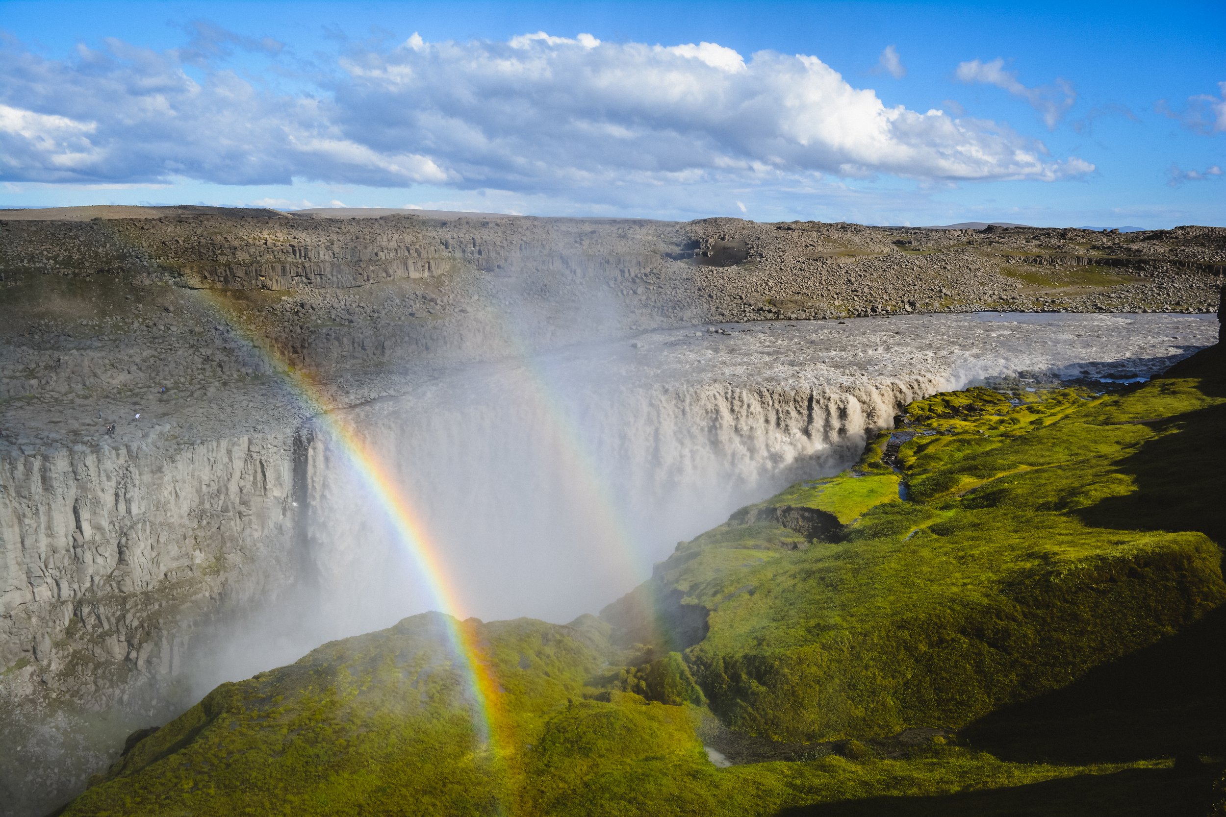 A waterfall with a rainbow arching over it, surrounded by green moss and rocky terrain under a partly cloudy sky.