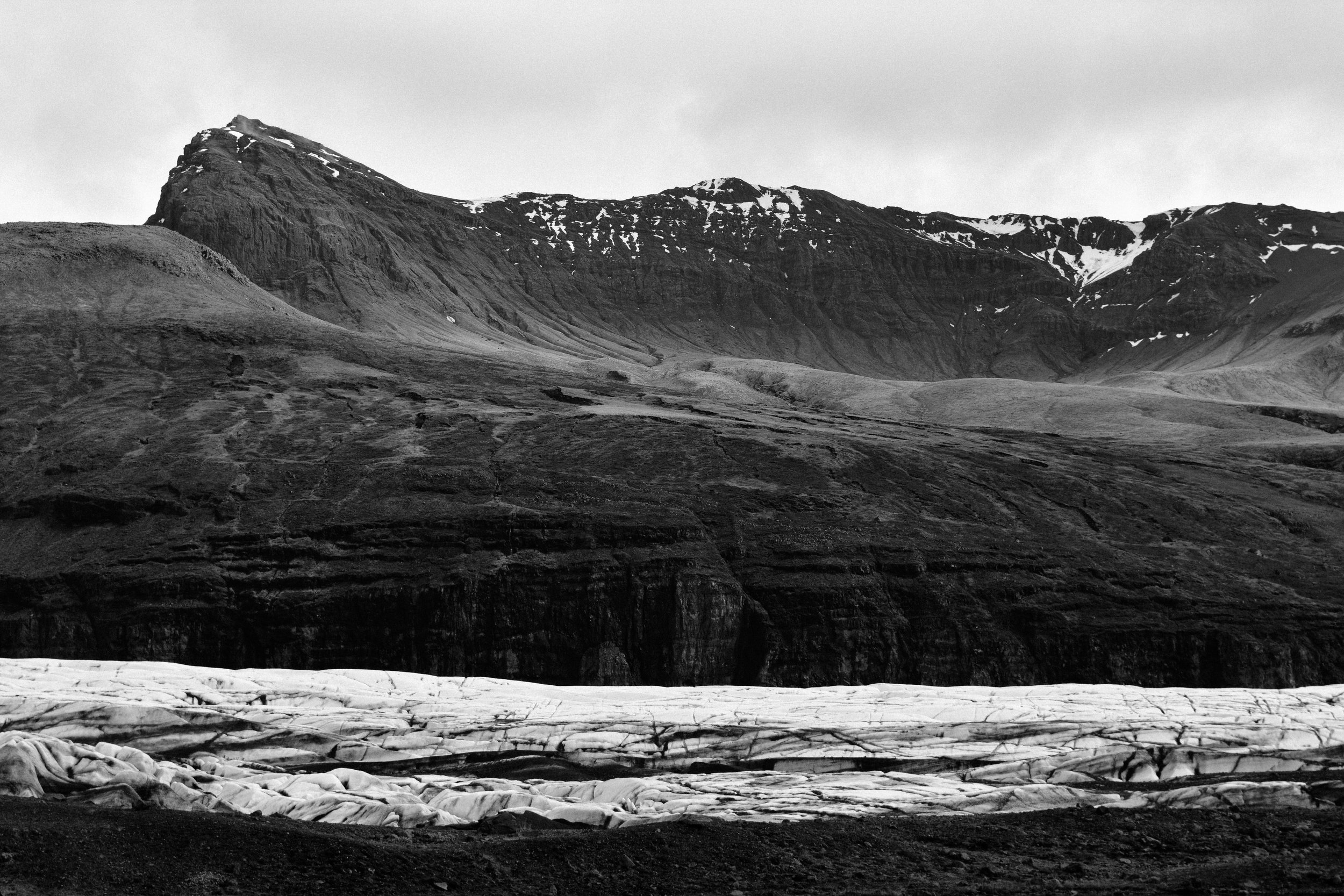 A black-and-white landscape of rugged mountainous terrain with snow patches, a glacier in the foreground, and cloudy sky.