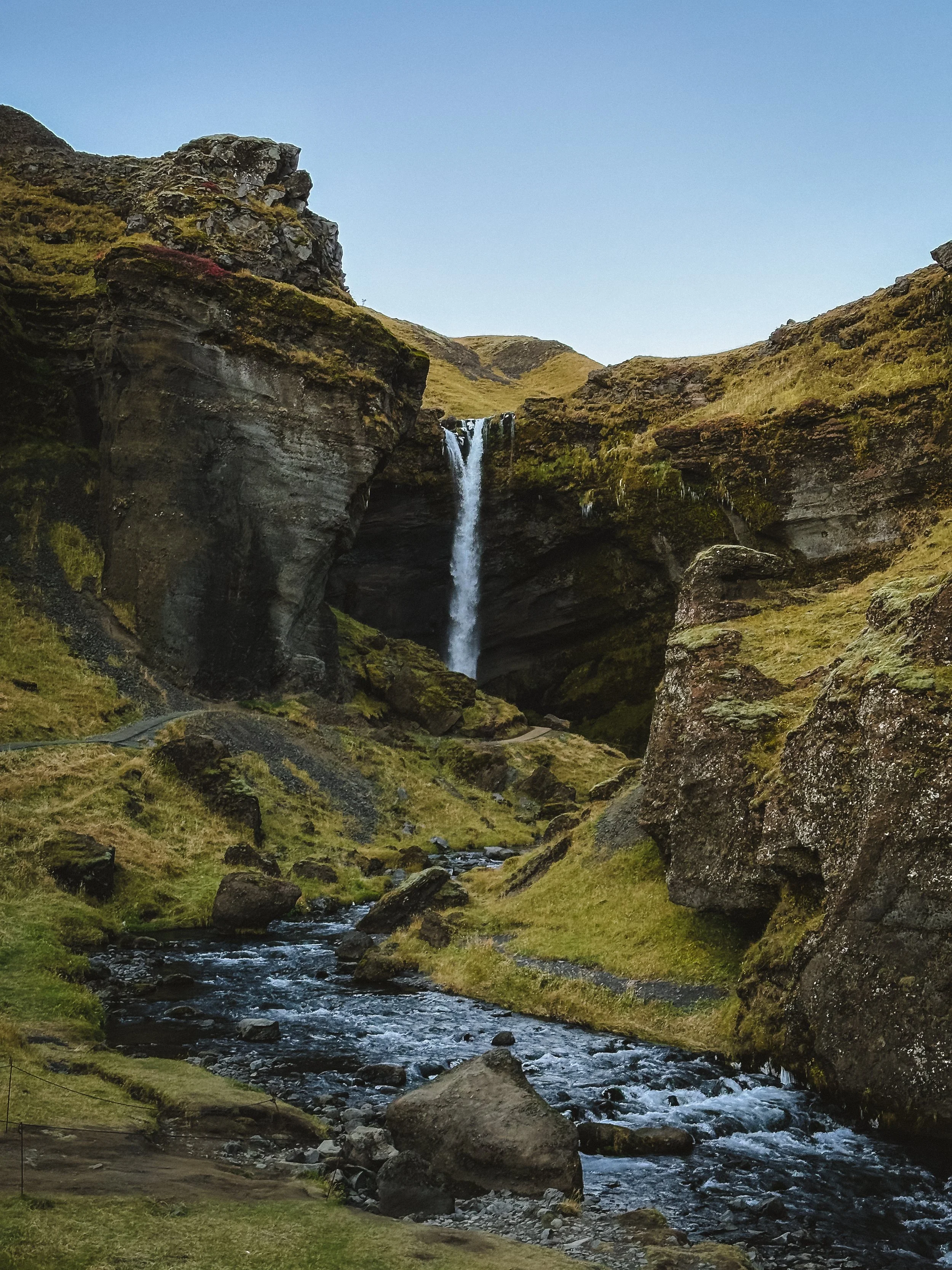A tall waterfall flowing down from a rocky cliff into a stream below, surrounded by moss-covered rocks and grassy terrain under a clear blue sky.