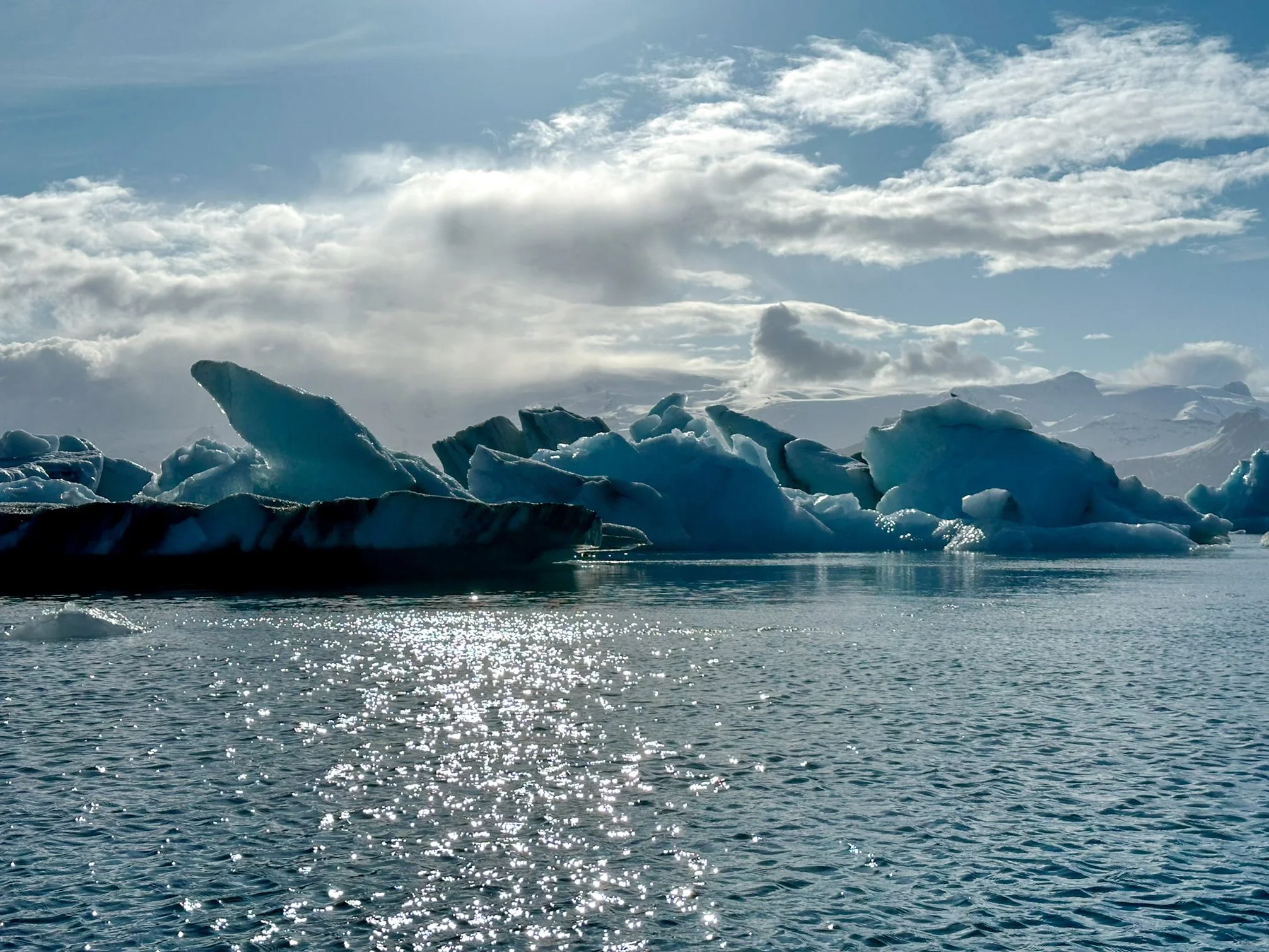 A glacier floating in a body of water with snow-capped mountains in the background and a cloudy sky.