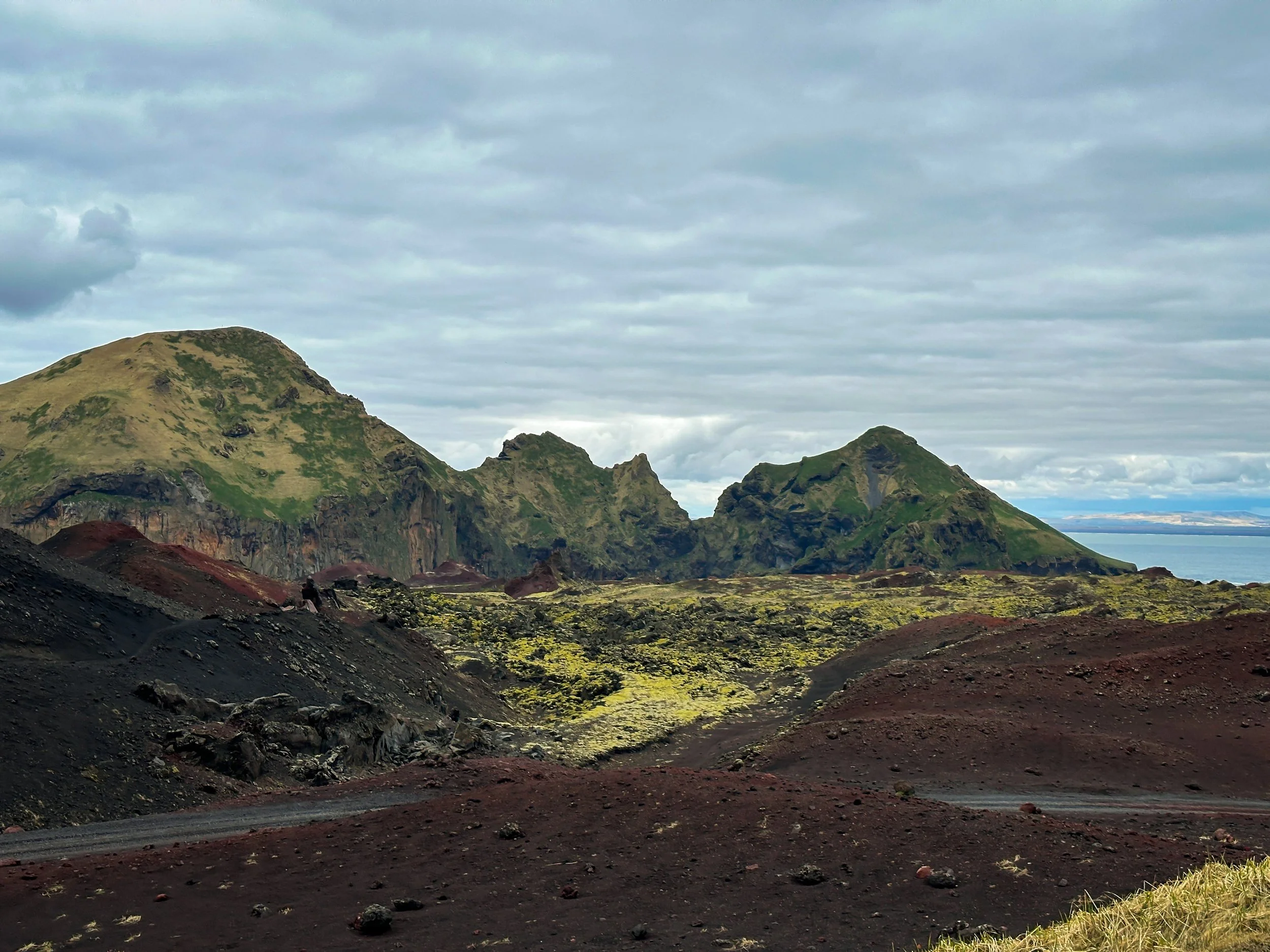 View of volcanic landscape with dark reddish-brown soil and green moss-covered rocky mountains in the background under a cloudy sky, with a distant body of water on the horizon.