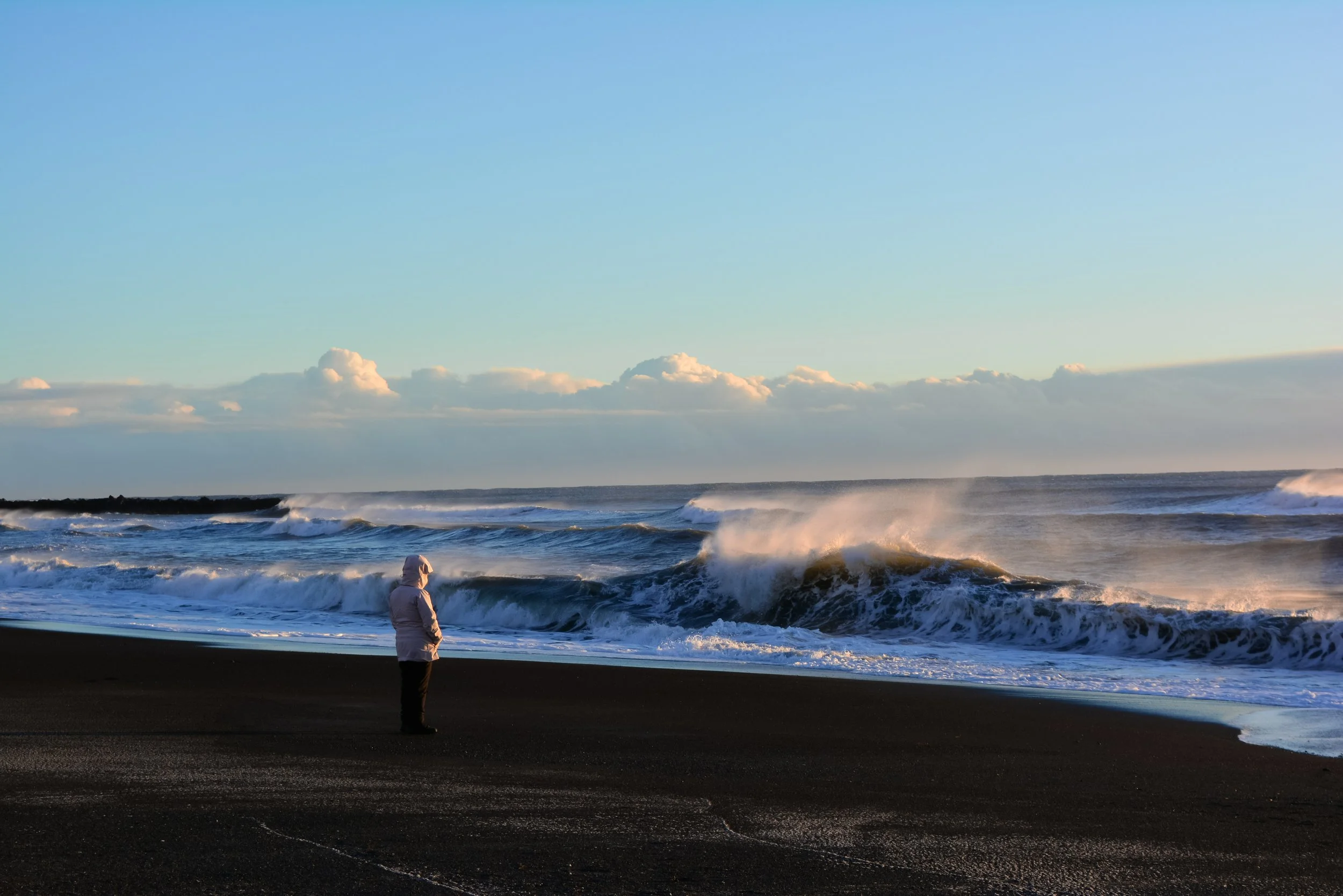 A person in a white jacket standing on a dark sandy beach looking at ocean waves during sunset or sunrise.