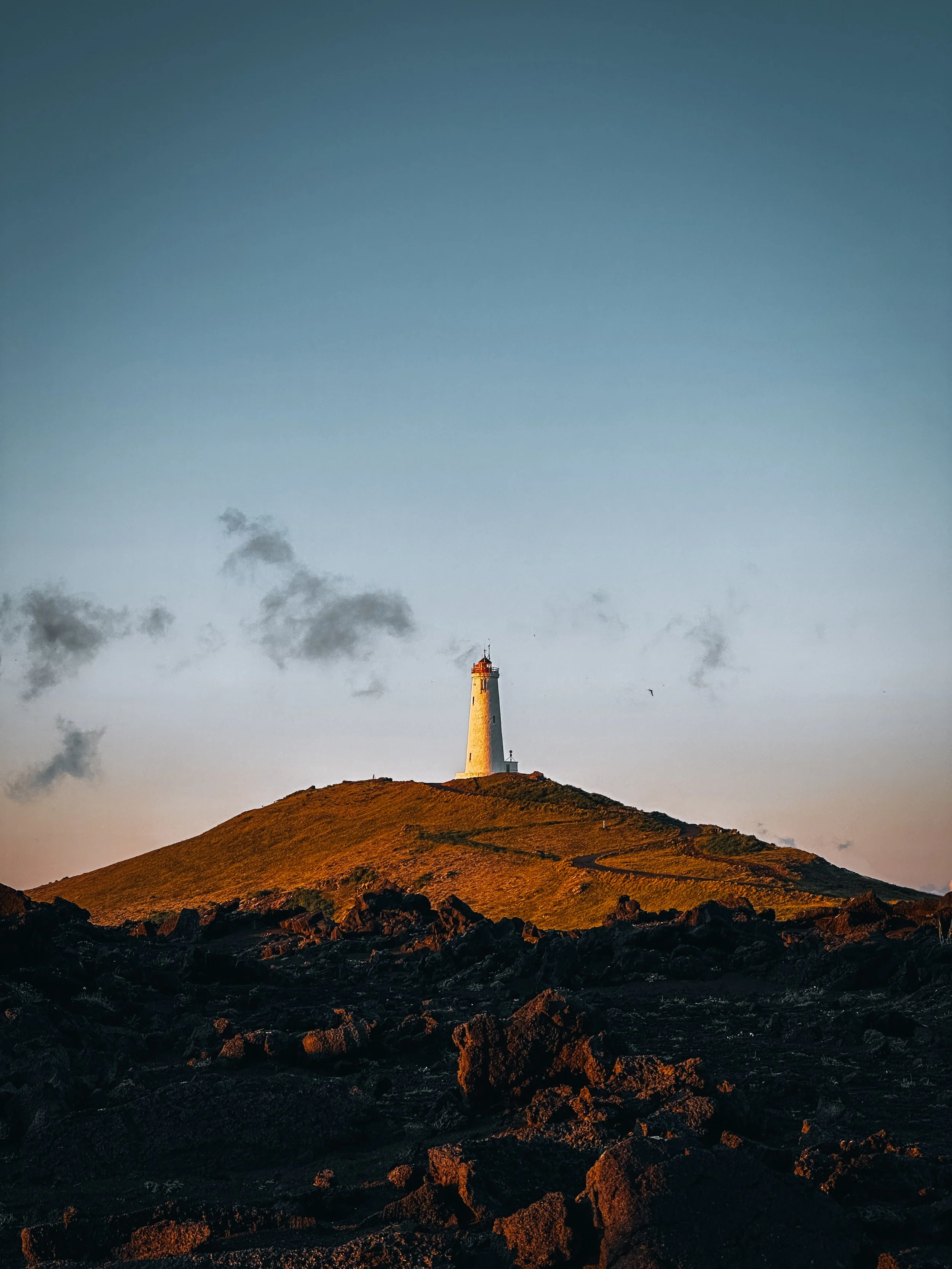 A lighthouse on a hilltop with rocky terrain in the foreground and a mostly clear sky with a few clouds in the background.