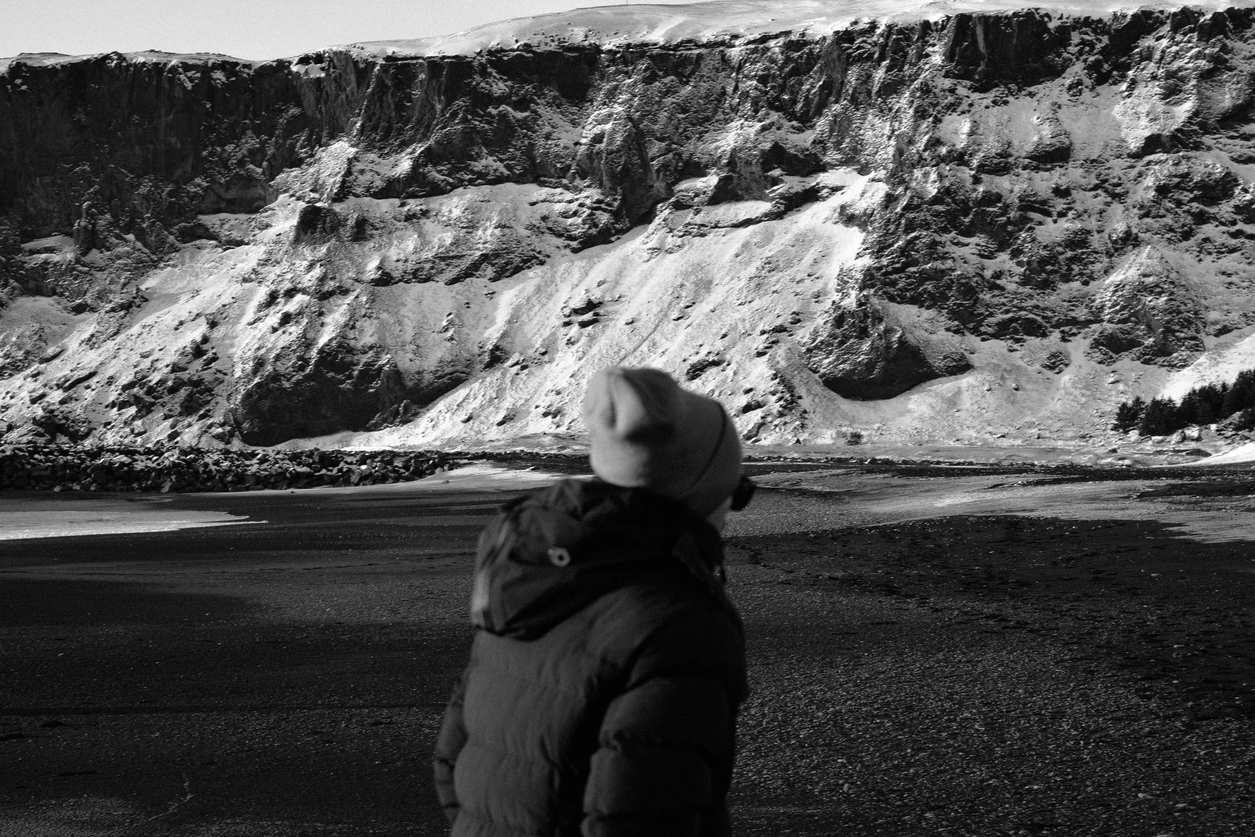 A person wearing a hat and winter coat looking at a snow-covered mountain with a body of water in the foreground.