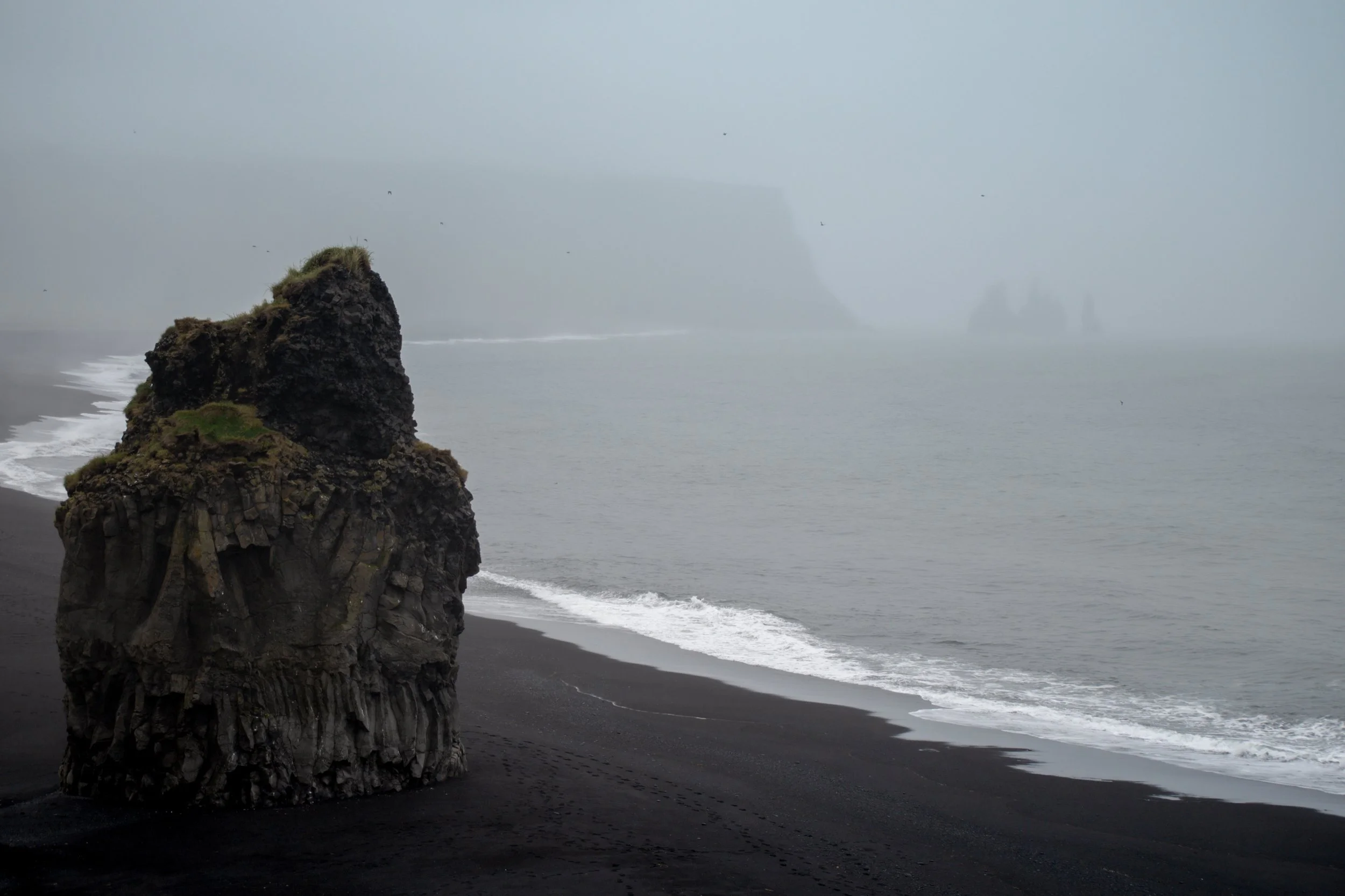 A large rock formation on a black sand beach with waves coming ashore. In the background, fog-shrouded cliffs or islands are visible, creating a moody and misty coastal scene.