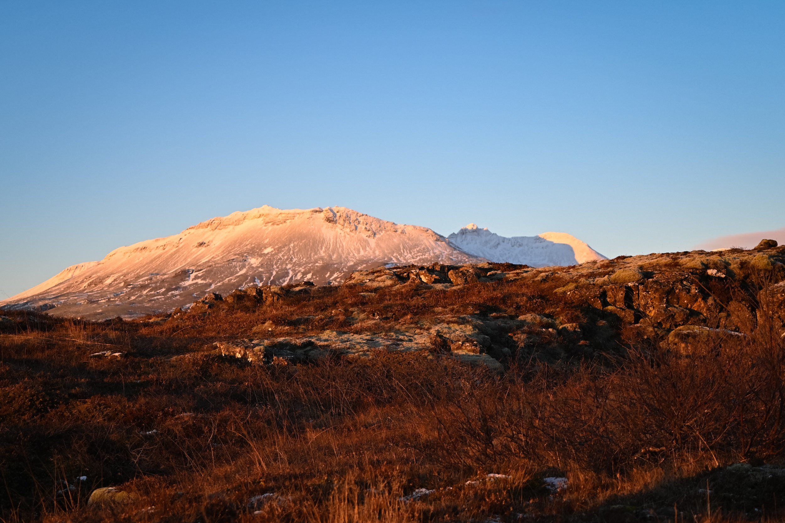 A landscape scene of snow-capped mountains in the distance with warm golden sunlight illuminating the rocky and shrub-covered foreground