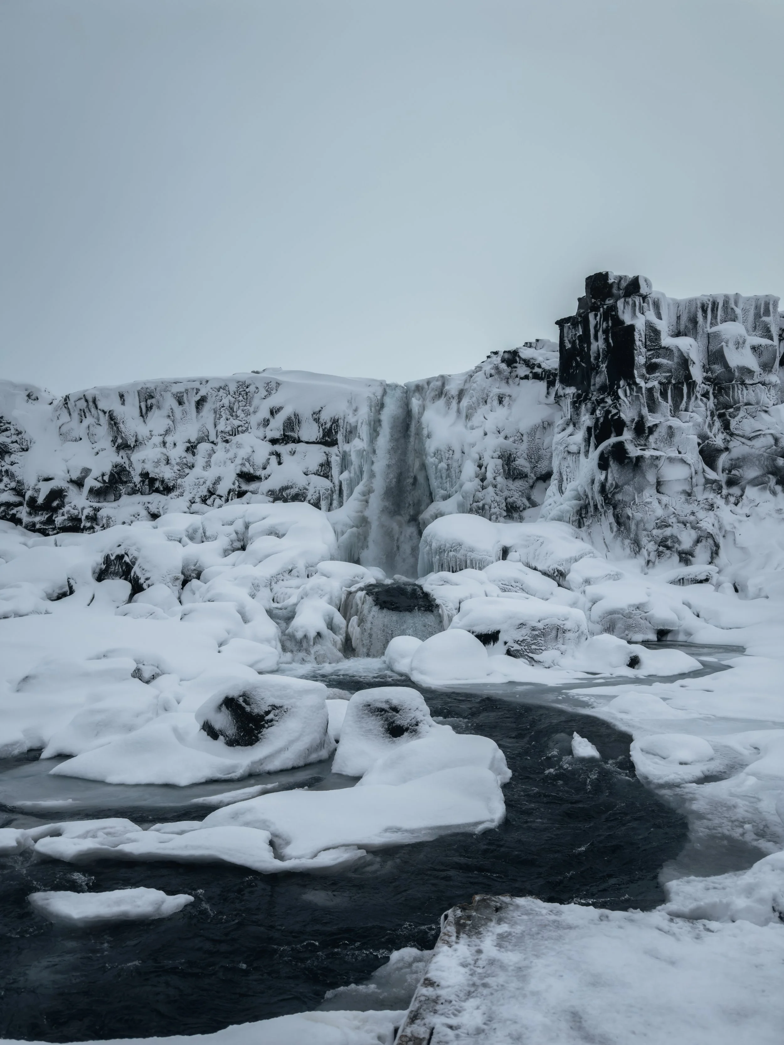 Frozen waterfall flowing into a partially frozen river surrounded by ice-covered rocks in a wintry landscape.