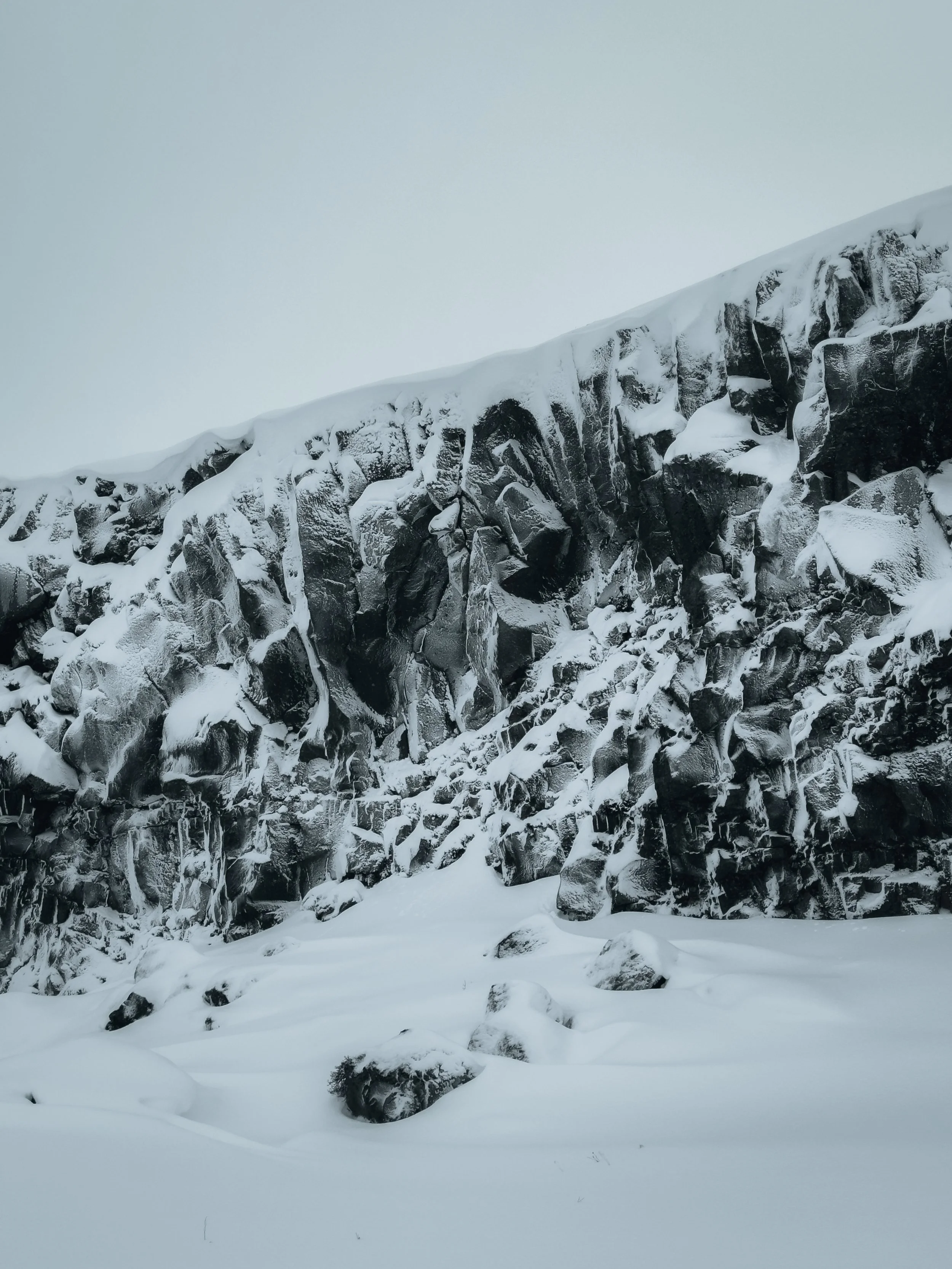 Snow-covered rocky cliff with icicles hanging from the rocks, under a cloudy sky.