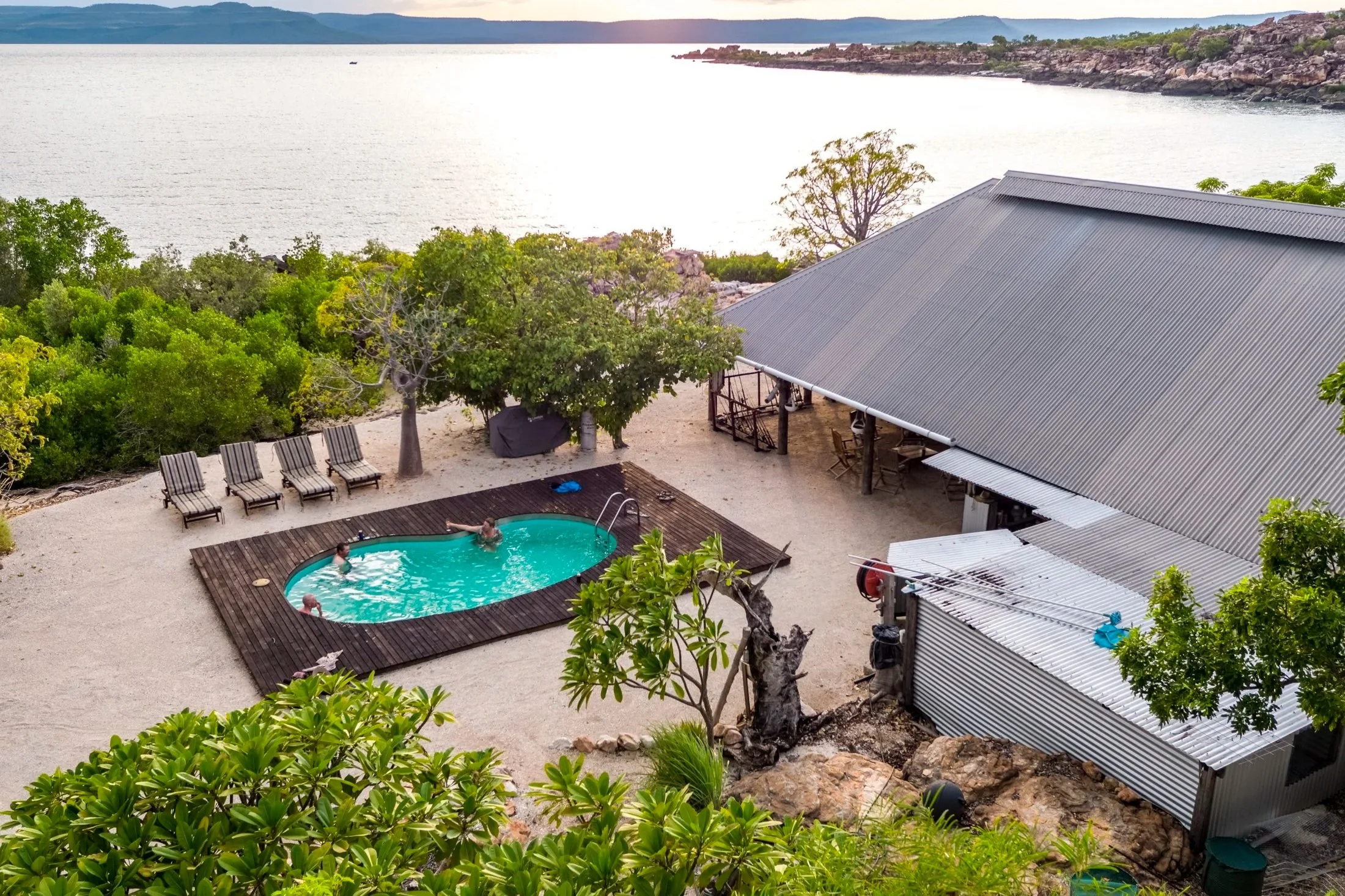 A scenic view of a backyard with a swimming pool, surrounded by lounge chairs, lush green trees, and a view of a large body of water in the background.
