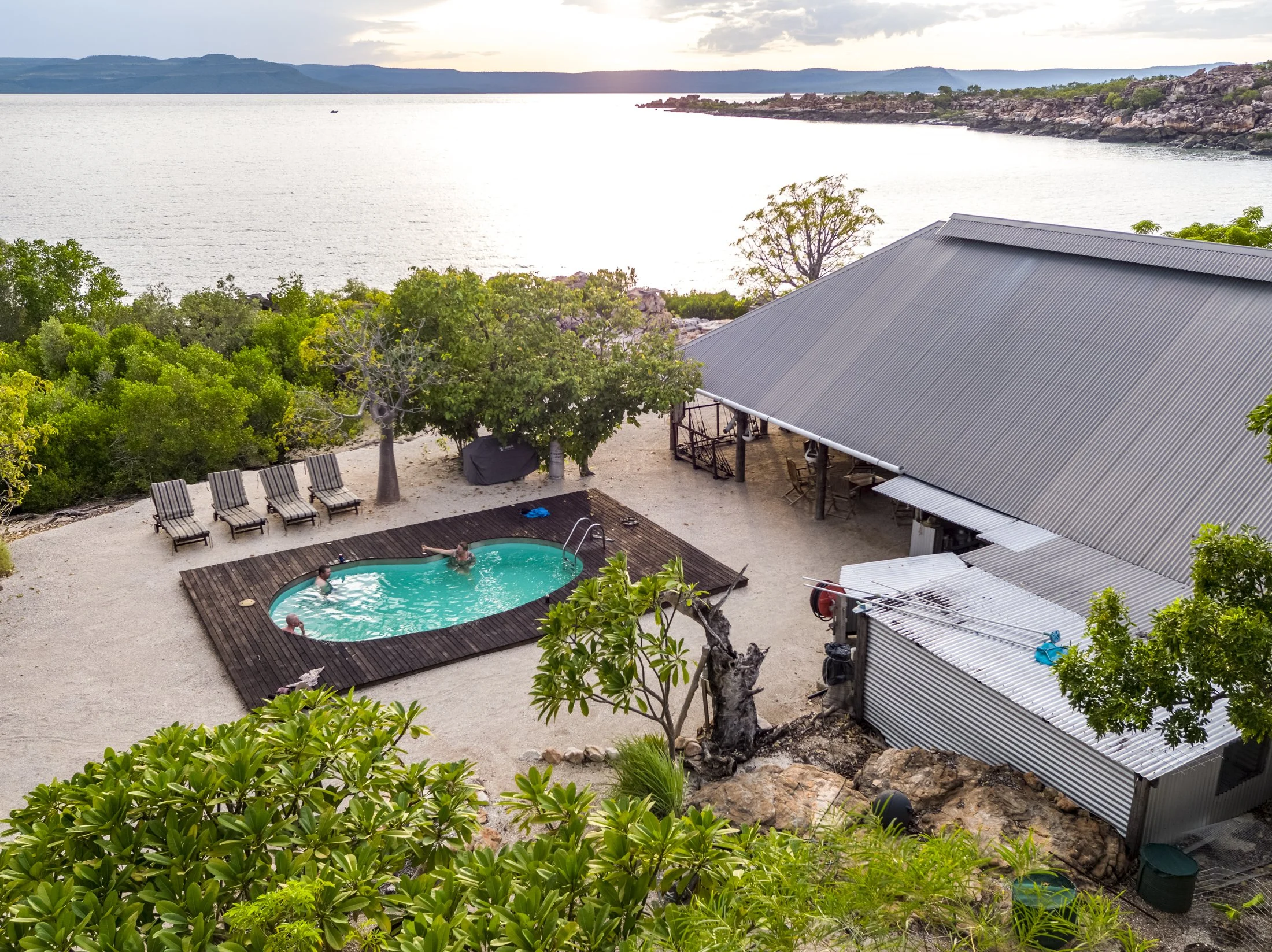 A backyard patio area by a lake with a small oval swimming pool, four lounge chairs, and trees surrounding the area. There is a large shed with a sloped metal roof and a deck, overlooking water and distant hills.