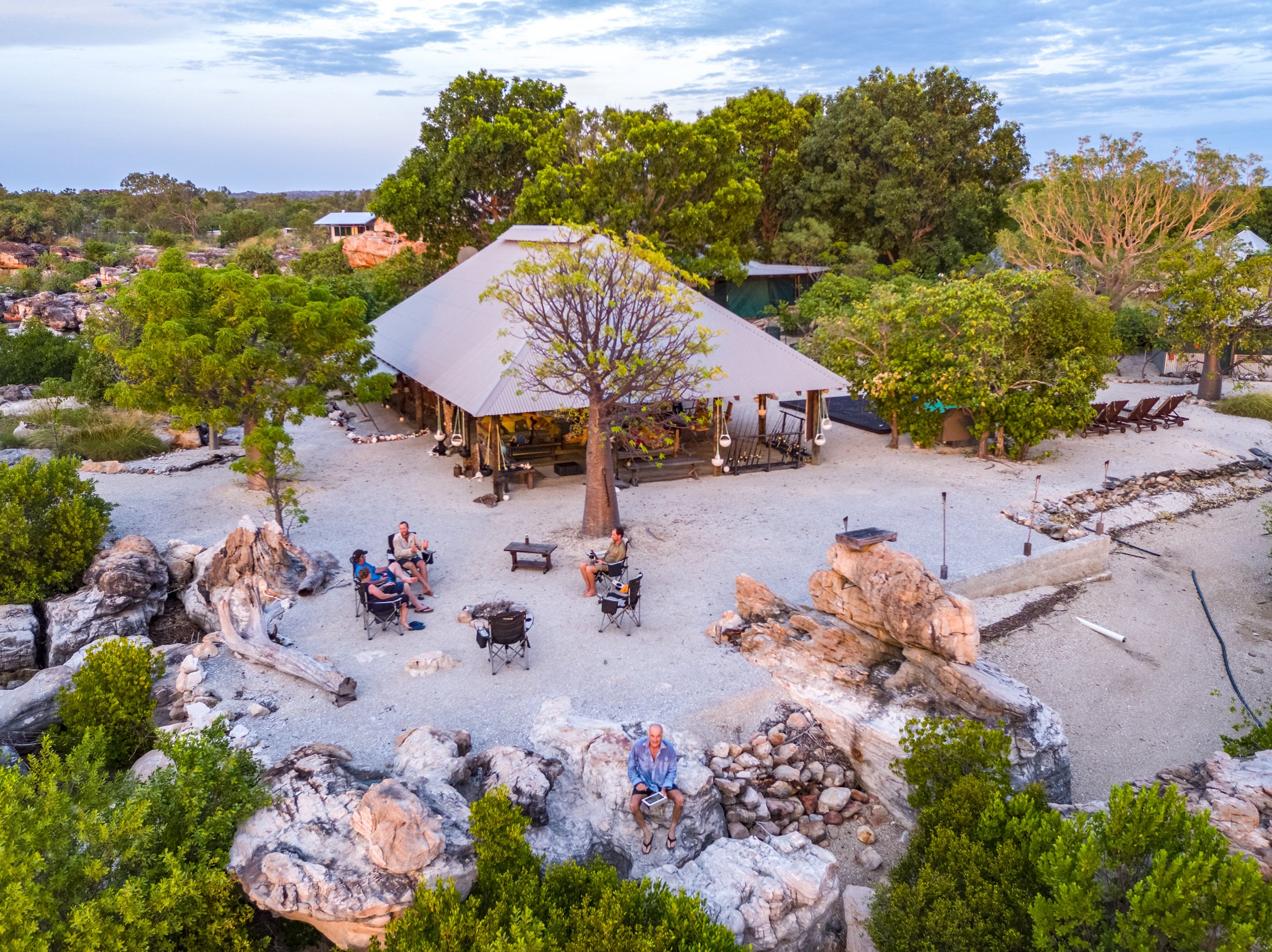 A backyard outdoor gathering area with a large metal-roofed pavilion, surrounded by trees, rocks, lounge chairs, and several people sitting around a fire pit, during sunset.