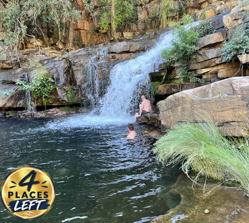 Two people swimming in a natural pool underneath a small waterfall with rocks and greenery surrounding the area.