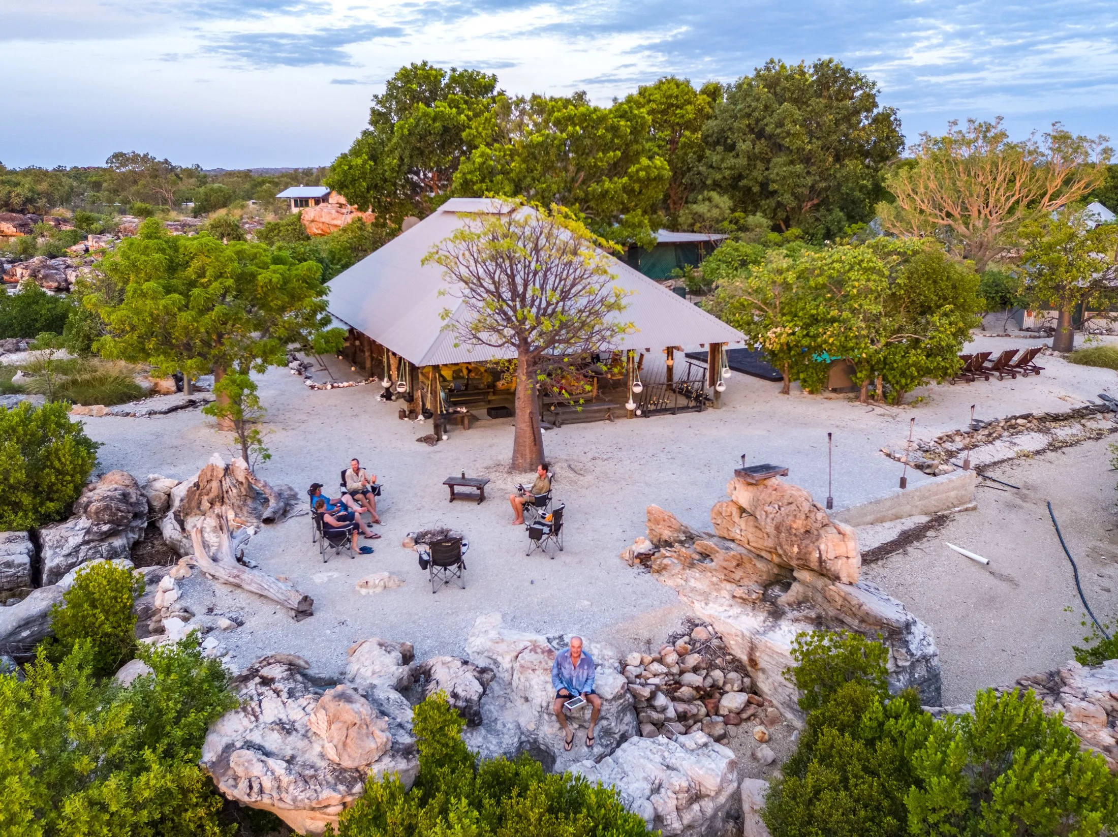 An outdoor gathering area with a large metal-roofed pavilion, surrounded by trees, rocks, and gravel. Several people are sitting in chairs around a firepit, engaging in conversation. There are lounge chairs on the right and a man sitting on rocks near the foreground. The setting appears to be a natural, scenic outdoor space, possibly at a retreat or resort.