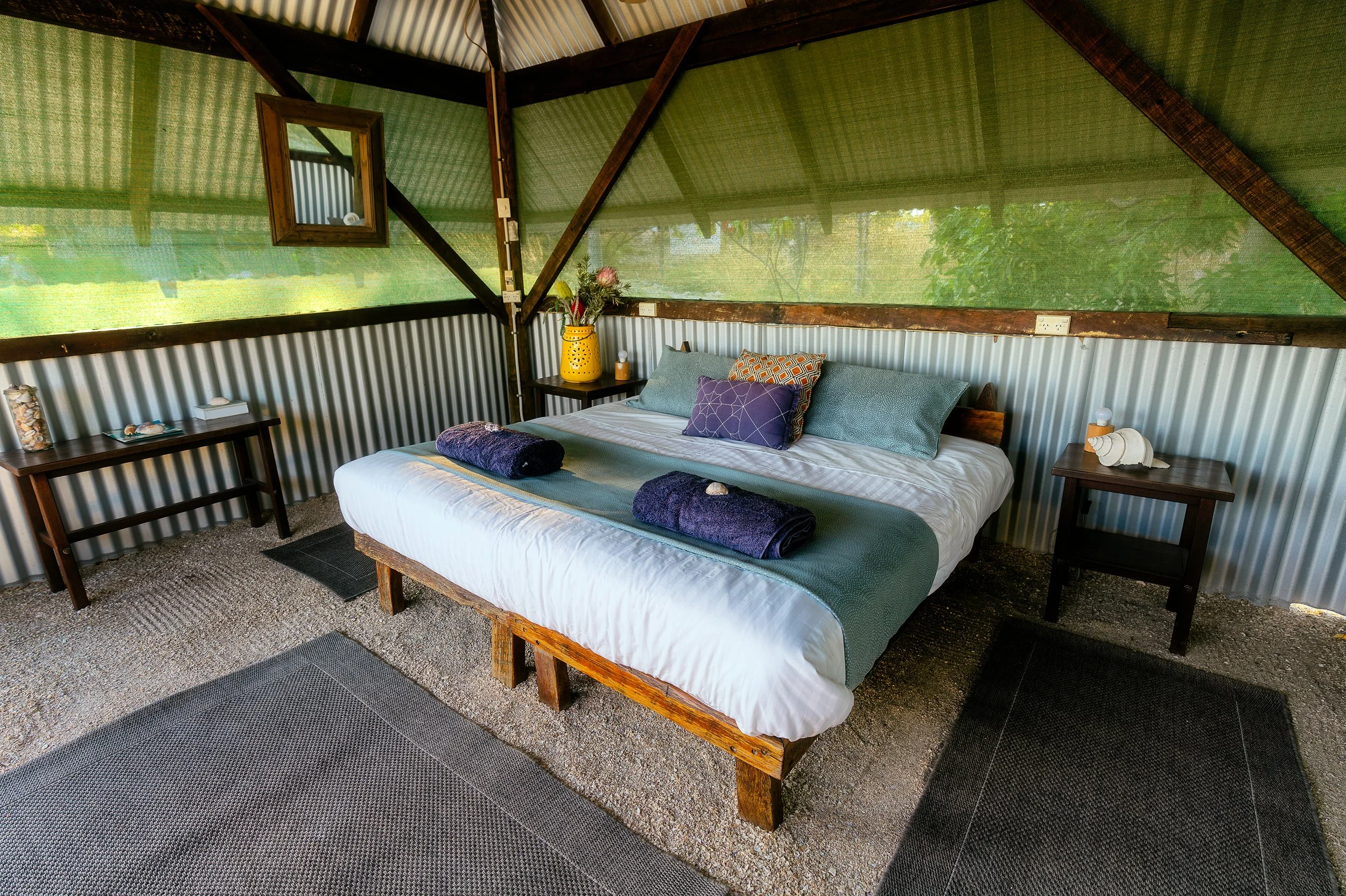 Interior of a rustic bedroom with a large bed, decorative pillows, rolled towels, wooden furniture, and green mesh walls allowing sunlight to filter through.