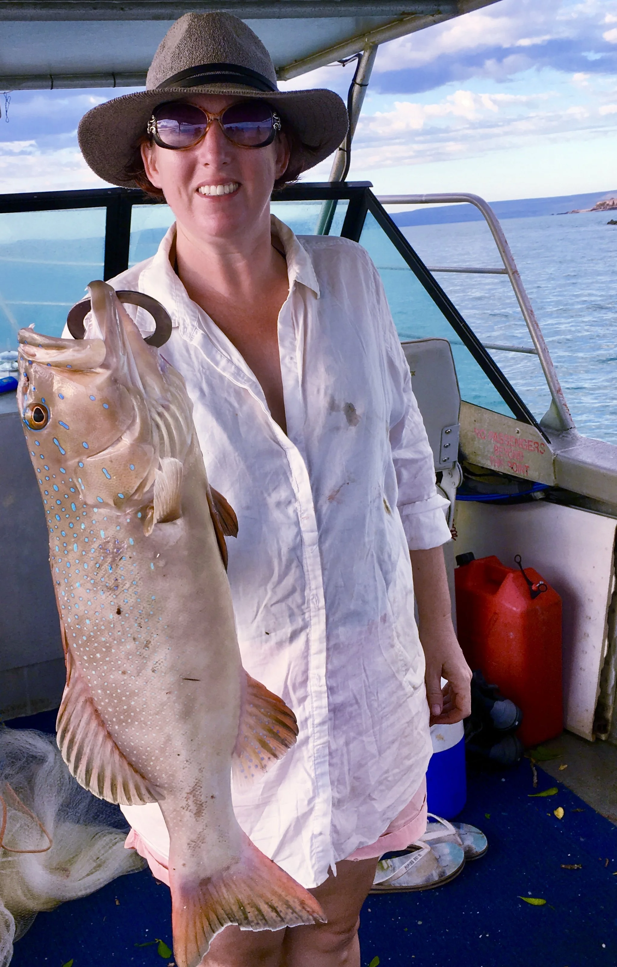 A woman wearing a sunhat and sunglasses holding a large fish on a boat with ocean and sky in the background.