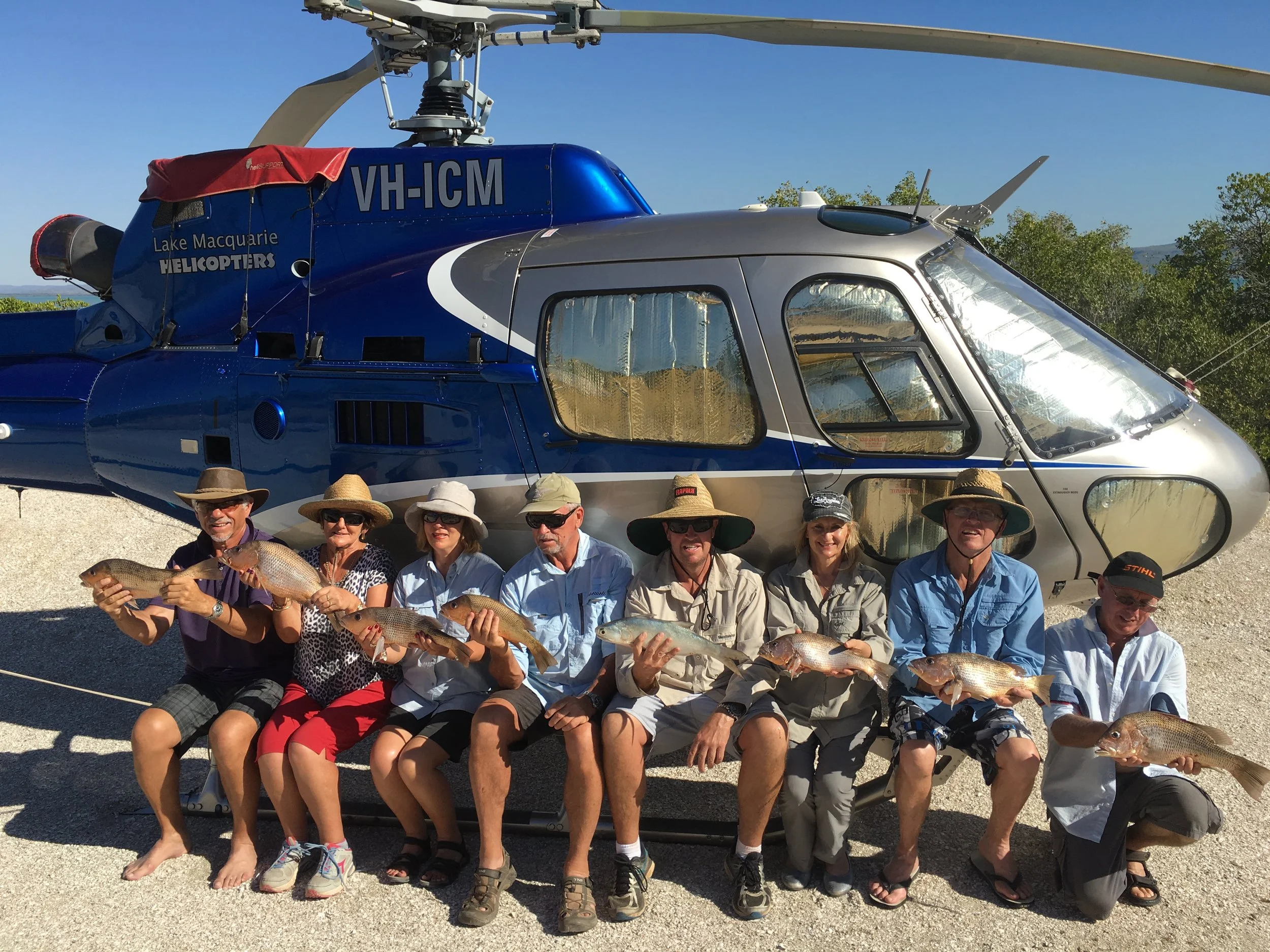 Group of people sitting in front of a helicopter holding caught fish, outdoors on a sunny day.