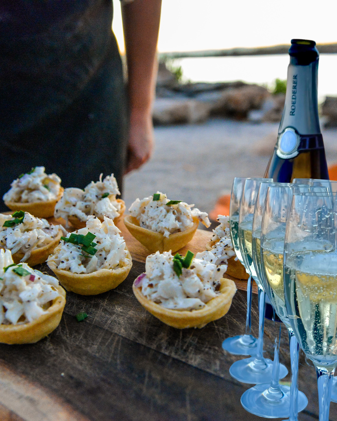 Appetizer bites with a creamy topping garnished with chopped green onions, set on a wooden platter alongside glasses of sparkling wine and a bottle of sparkling water.