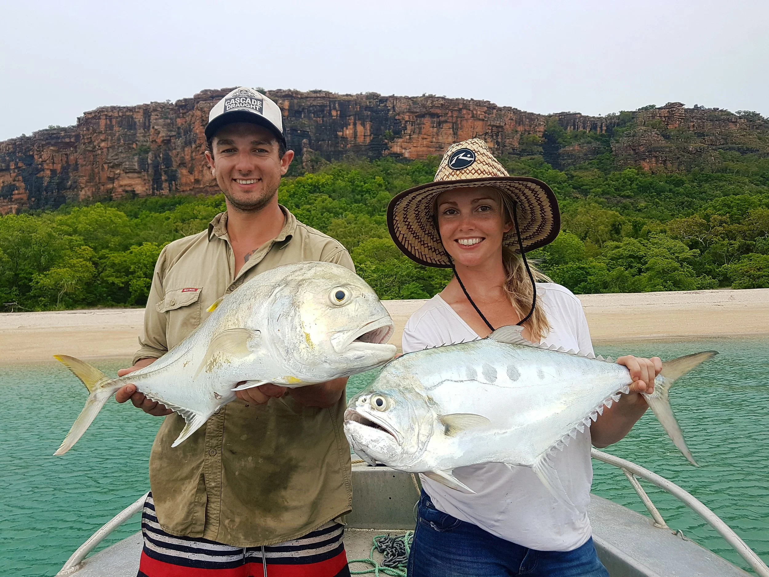 A young man and woman on a boat holding large fish with a tropical landscape of green trees and cliffs in the background.