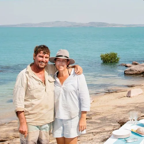 A man and woman smiling and posing together on a beach with water and hills in the background, with a table set with plates, glasses, and a blue tablecloth nearby.