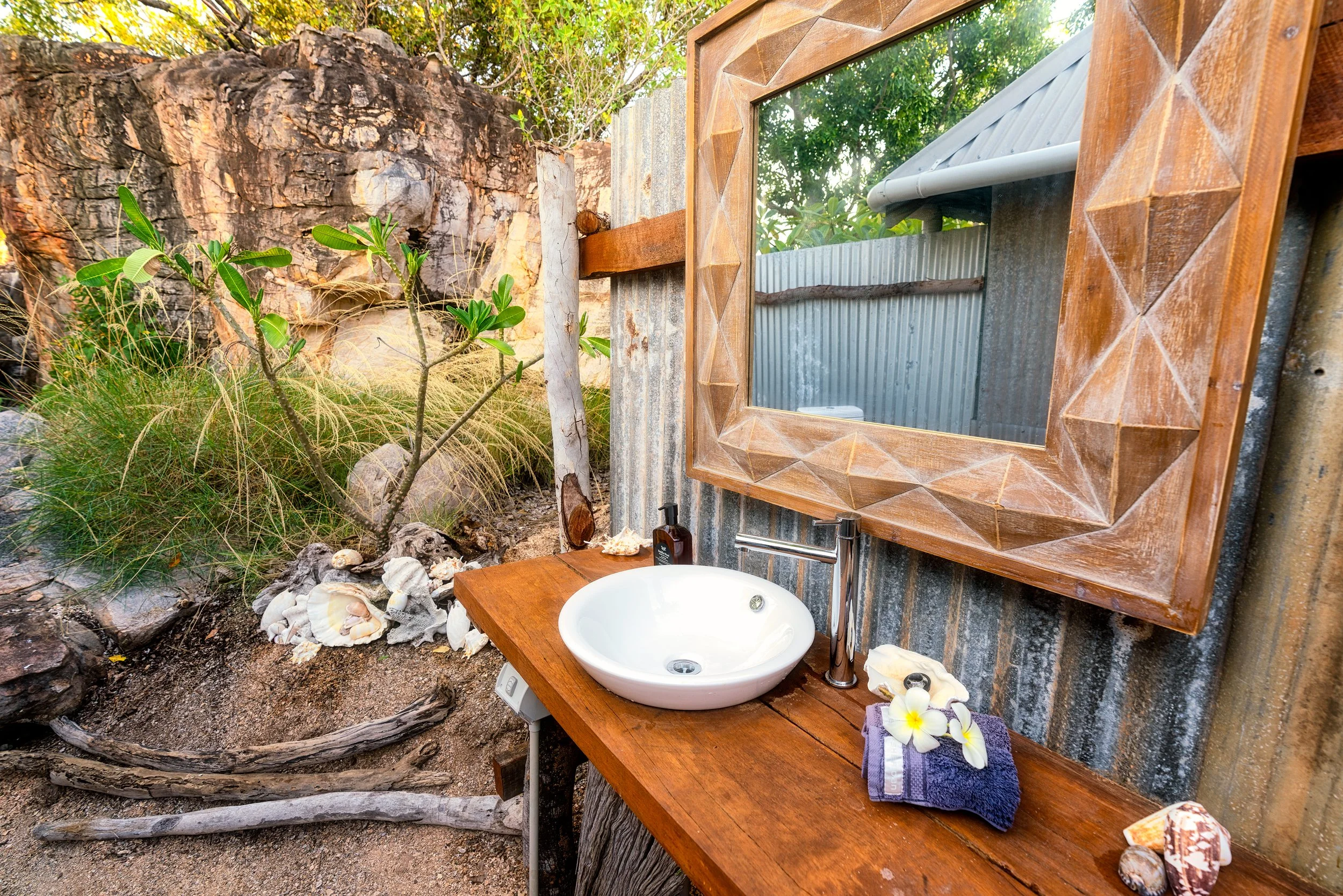 Outdoor bathroom sink with a wooden countertop, seashells, a towel with a flower, and a mirror, surrounded by rocks, plants, and a large tree trunk.