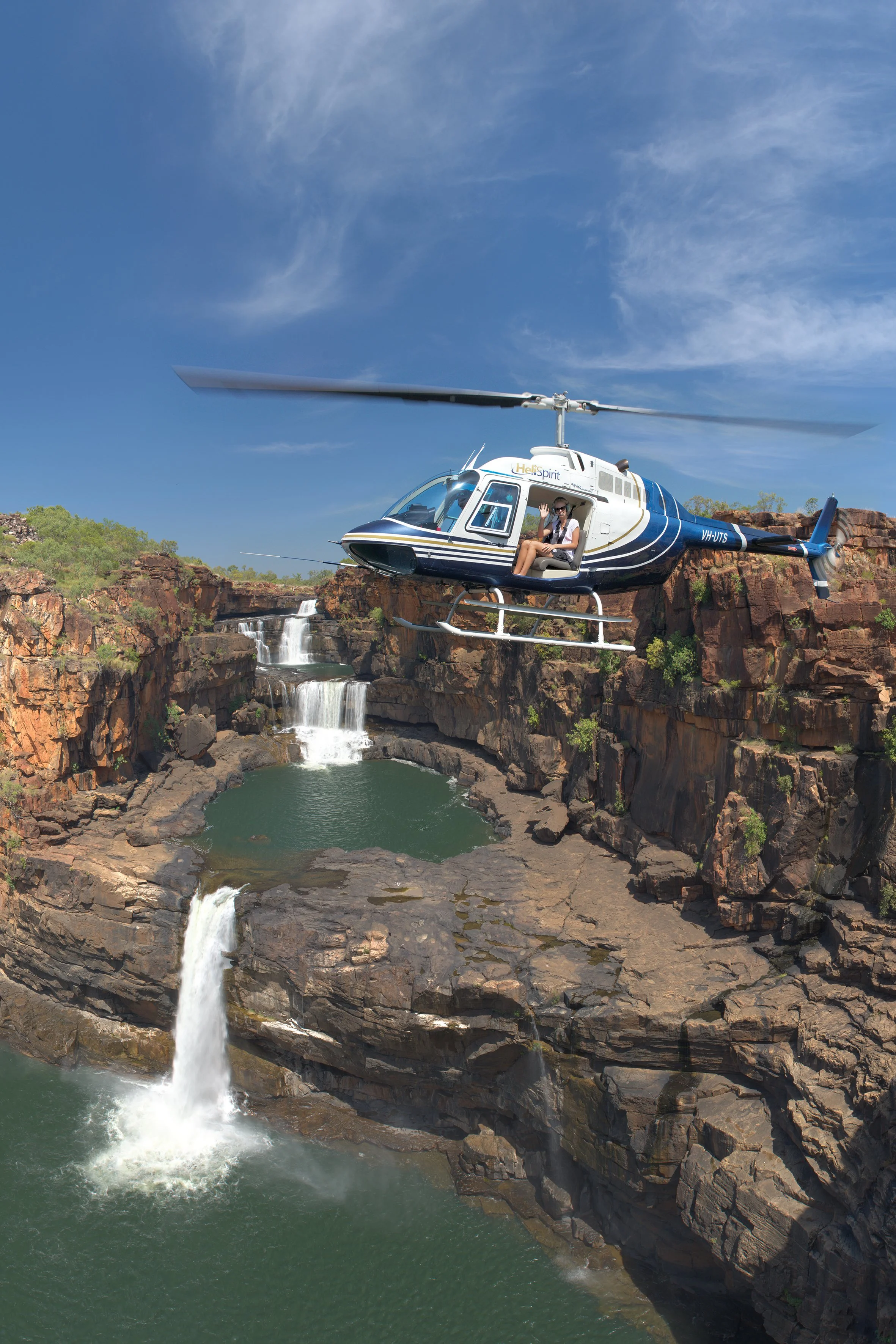A helicopter flying over a waterfall in a rocky canyon with greenery and blue sky in the background.