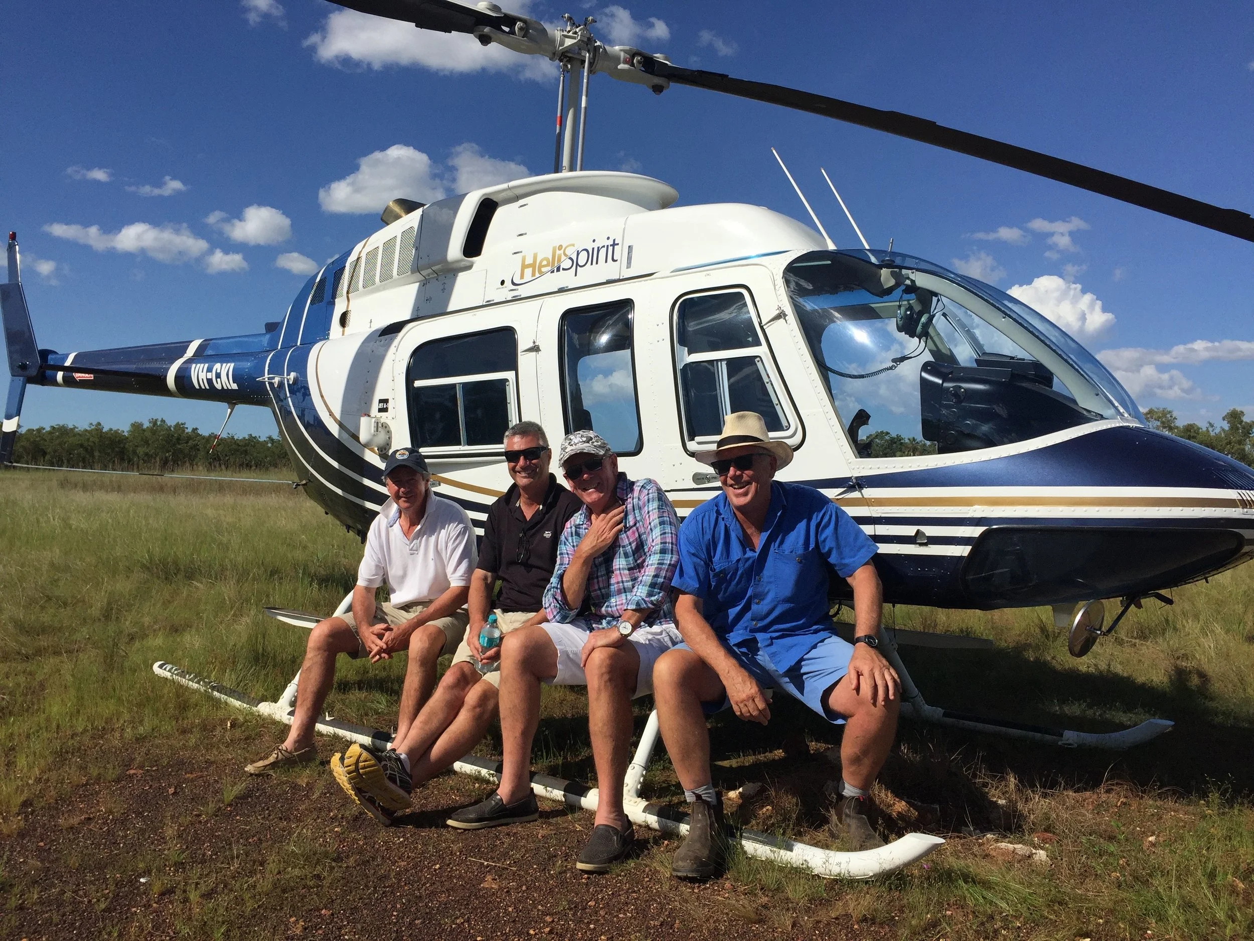 Four men sitting in front of a blue and white helicopter on a grassy field.