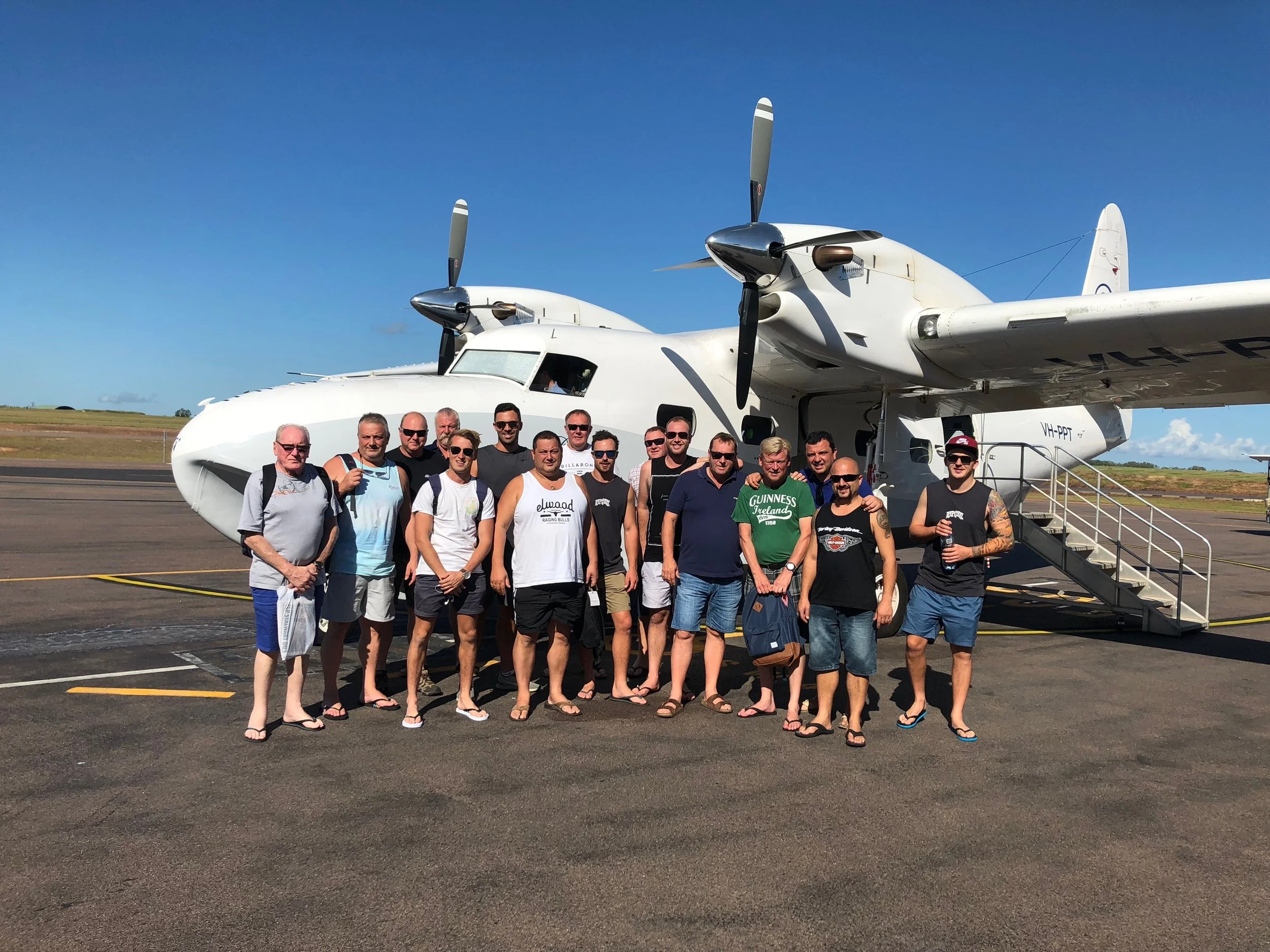 Group of people in casual clothing standing in front of a white twin-engine airplane on an airport tarmac on a sunny day.