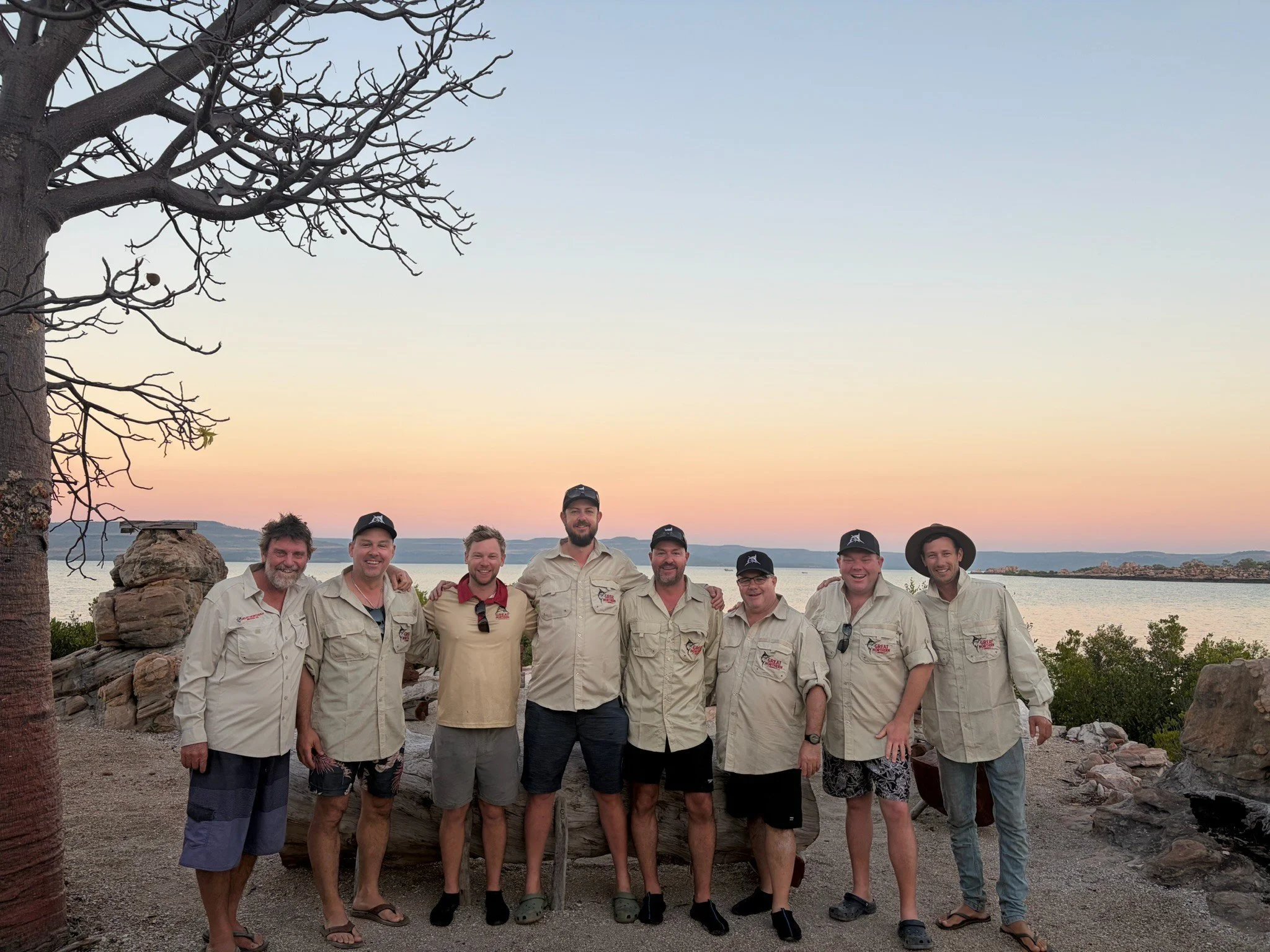 A group of ten smiling men standing outdoors at sunset, with a body of water and hills in the background, and a tree on the left side of the image.