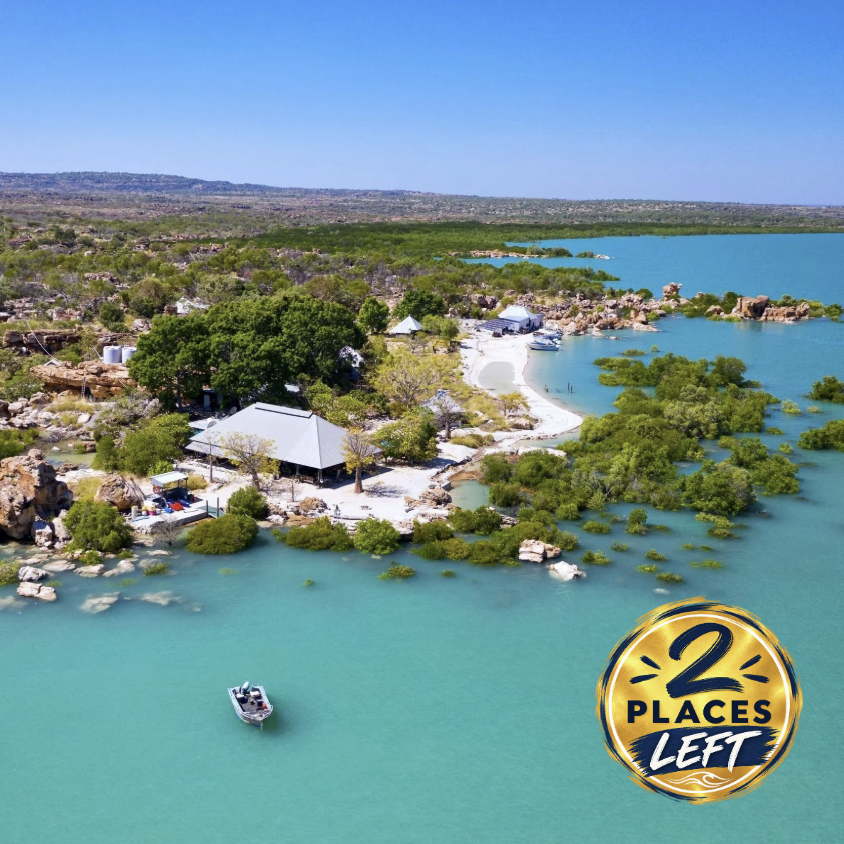 Aerial view of a lakeside area with a large building, trees, small boats, and a sandy beach, with a vast blue lake and distant landscape in the background during daytime.