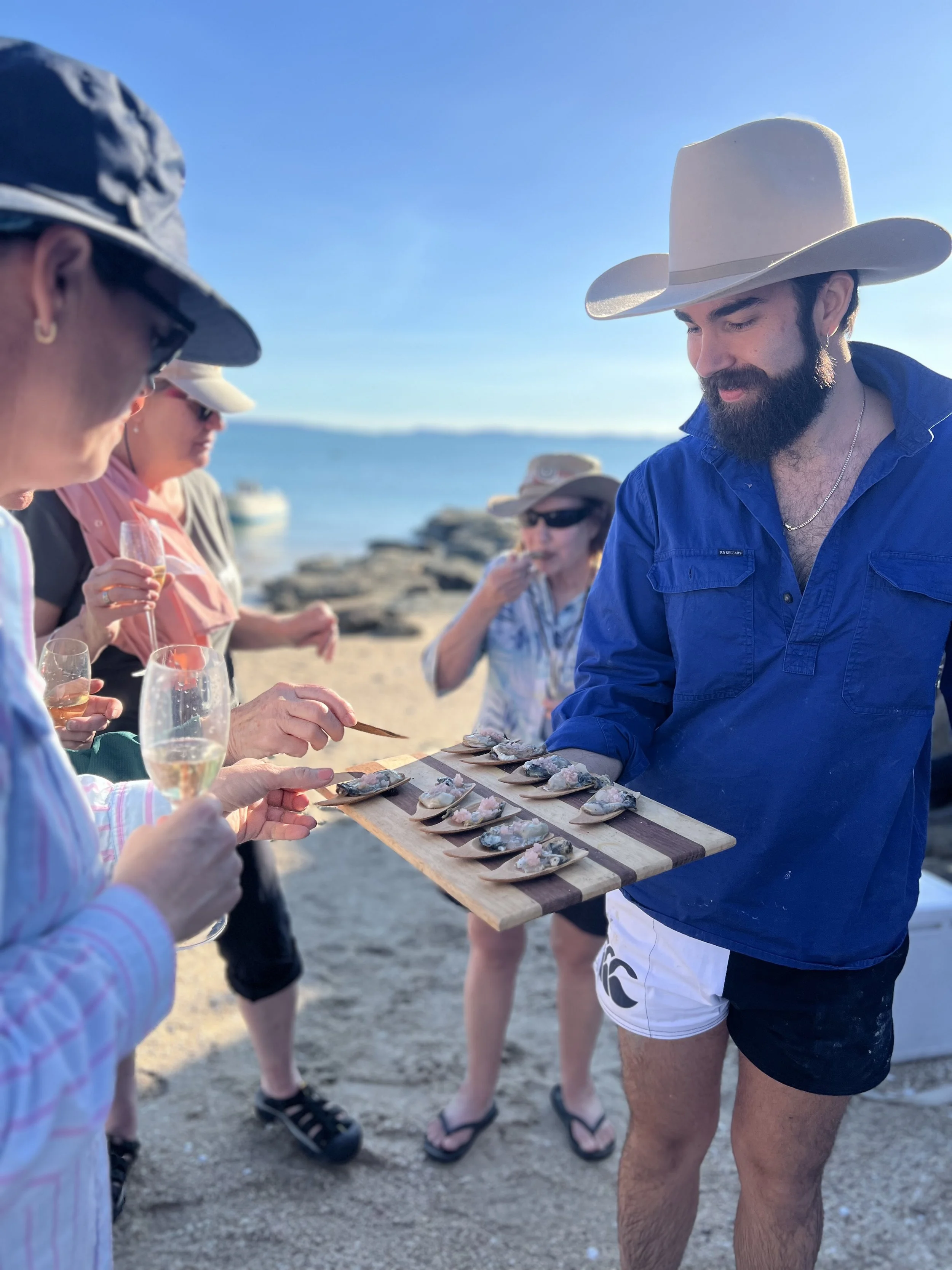 Group of people at the beach enjoying seafood and drinks, with a man offering small seafood dishes on a wooden board to others.