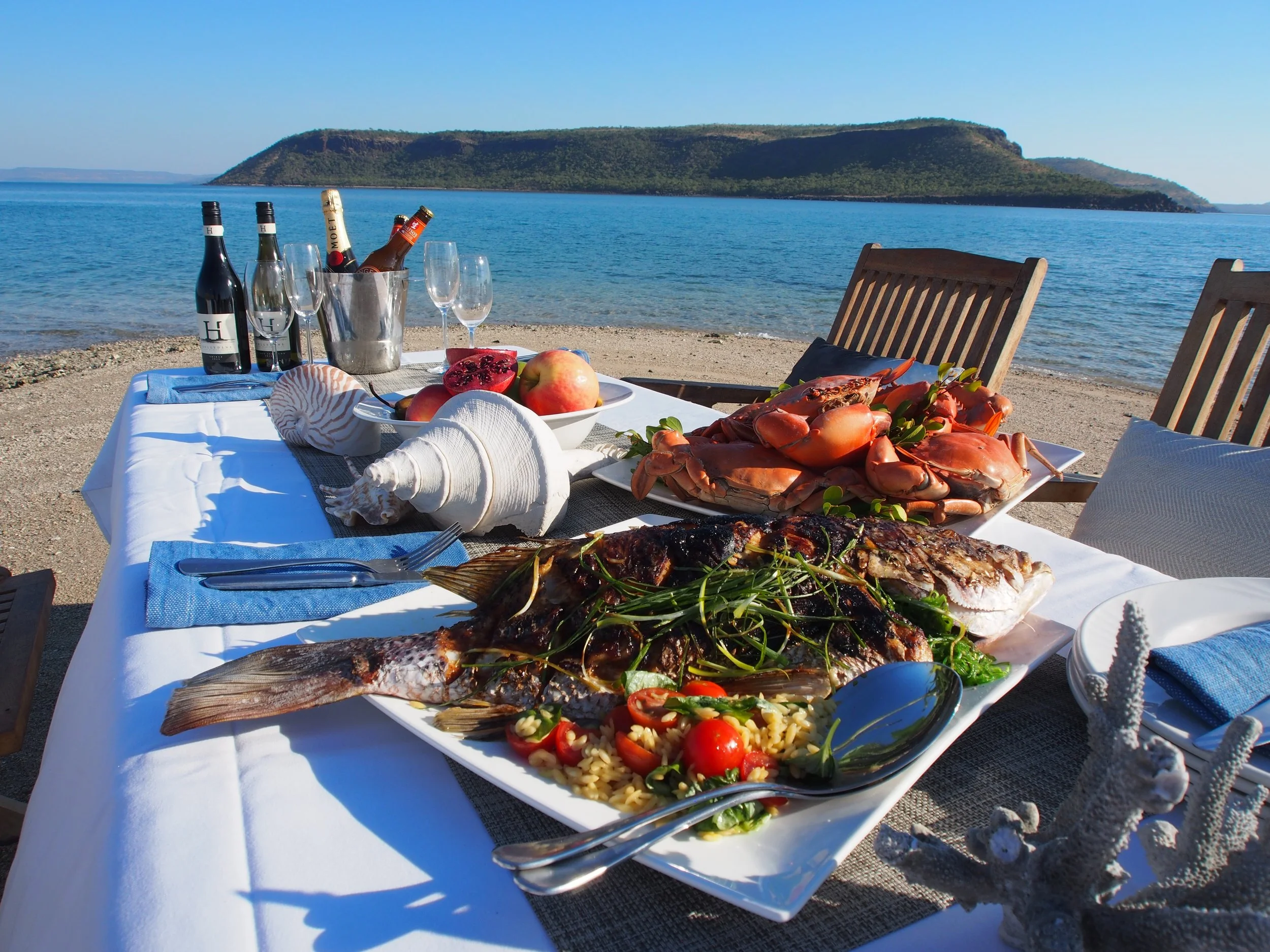 A seafood feast on a table by the beach, featuring grilled fish, crabs, rice with vegetables, and a shellfish arrangement, with wine, champagne, and fruit, against a backdrop of water and hills.