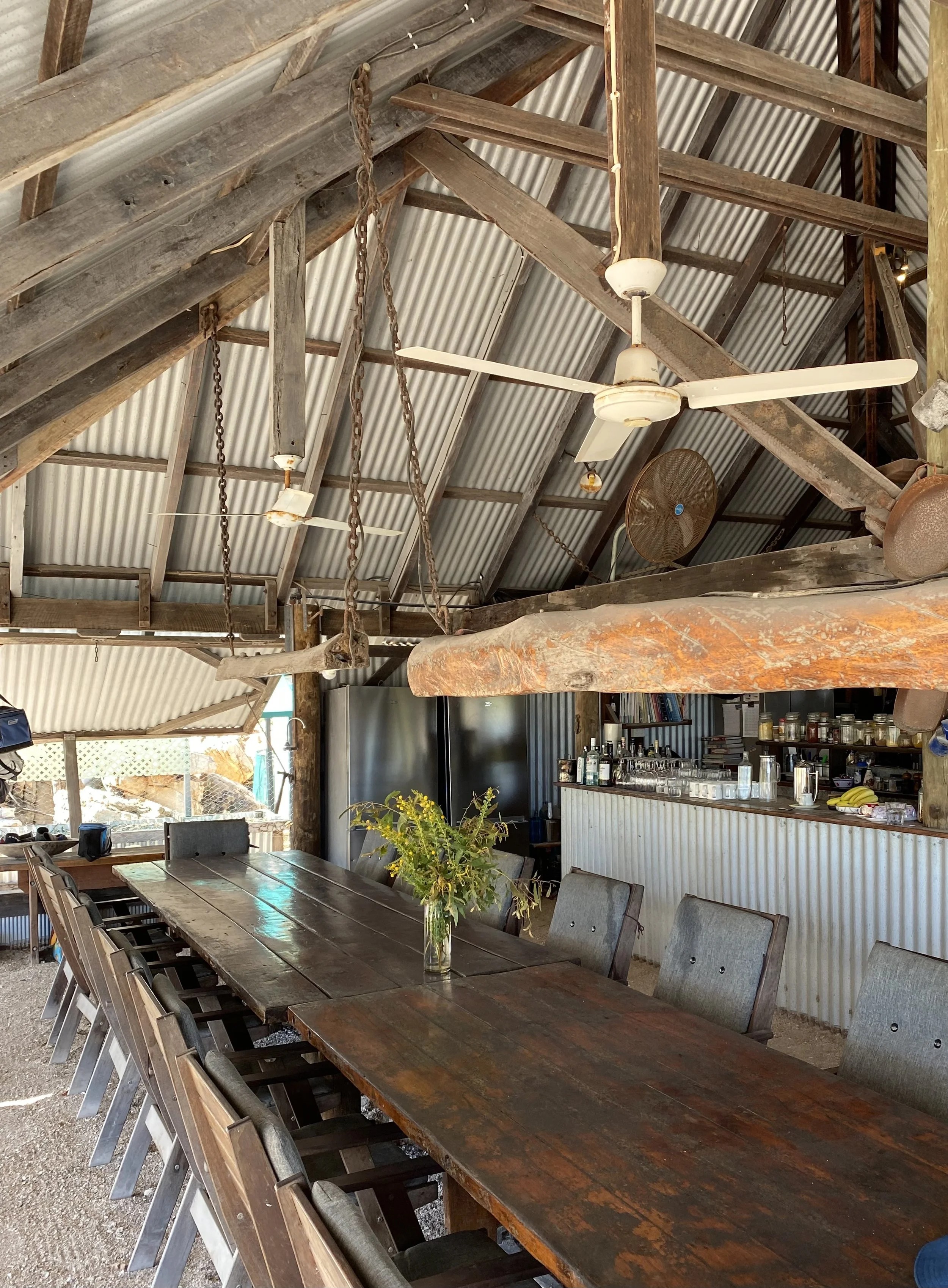 Rustic dining area with built-in long wooden table, surrounding chairs, and a bar in the background with various bottles, glasses, and a vase with flowers on the table, under a metal and wood roof with ceiling fans.