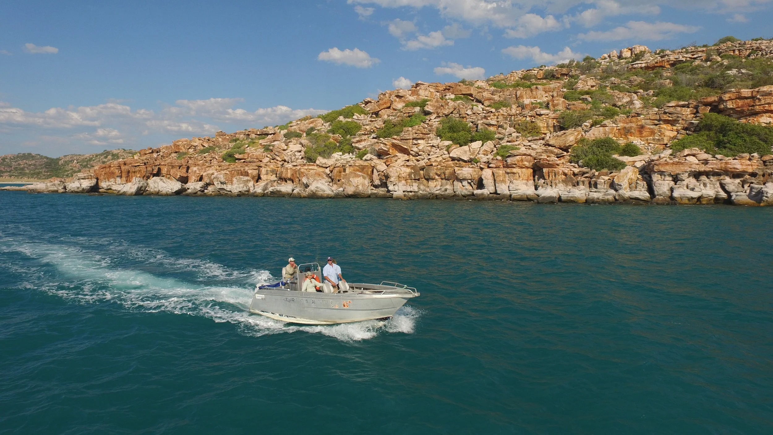 A small boat with three people aboard navigating a body of water near a rocky shoreline with sparse green vegetation under a partly cloudy sky.