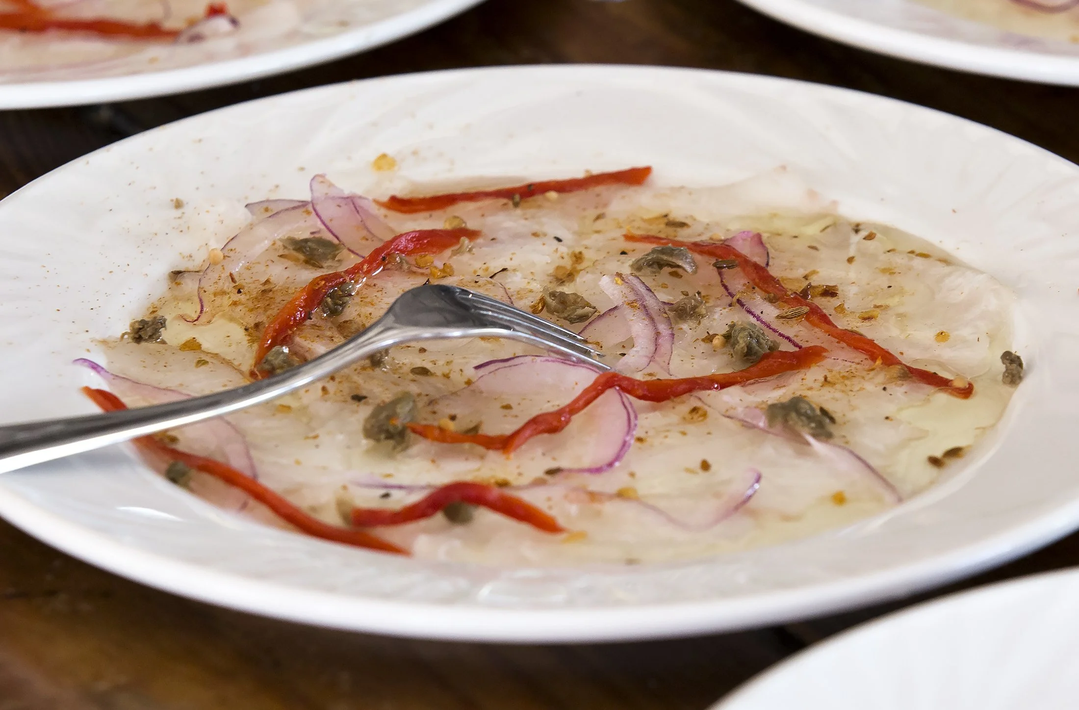 A white plate with ceviche garnished with red chili slices, red onion slices, and seeds, accompanied by a metal spoon.