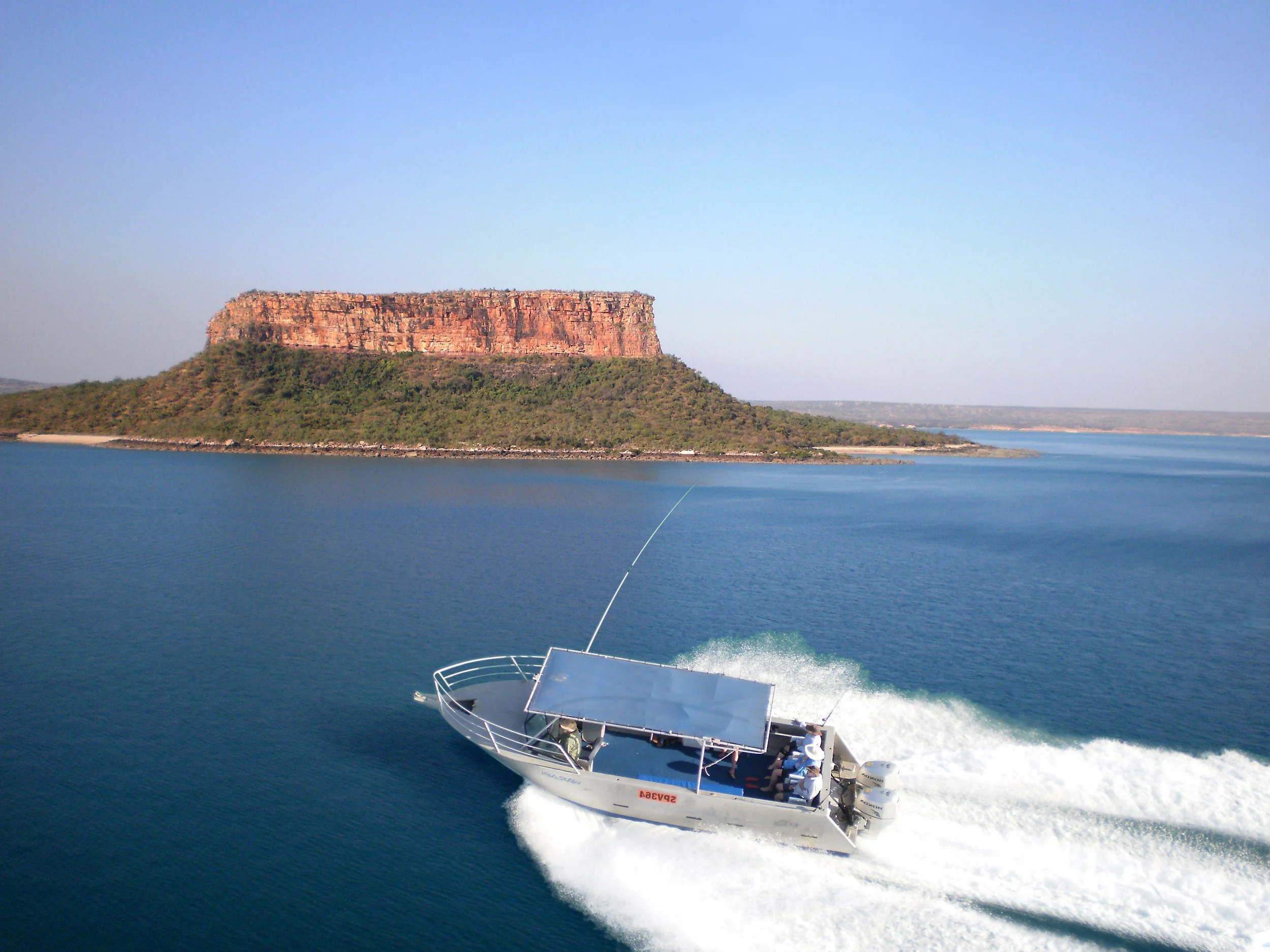 A boat with a solar panel on top speeding across a large body of water with a flat-topped hill in the background.