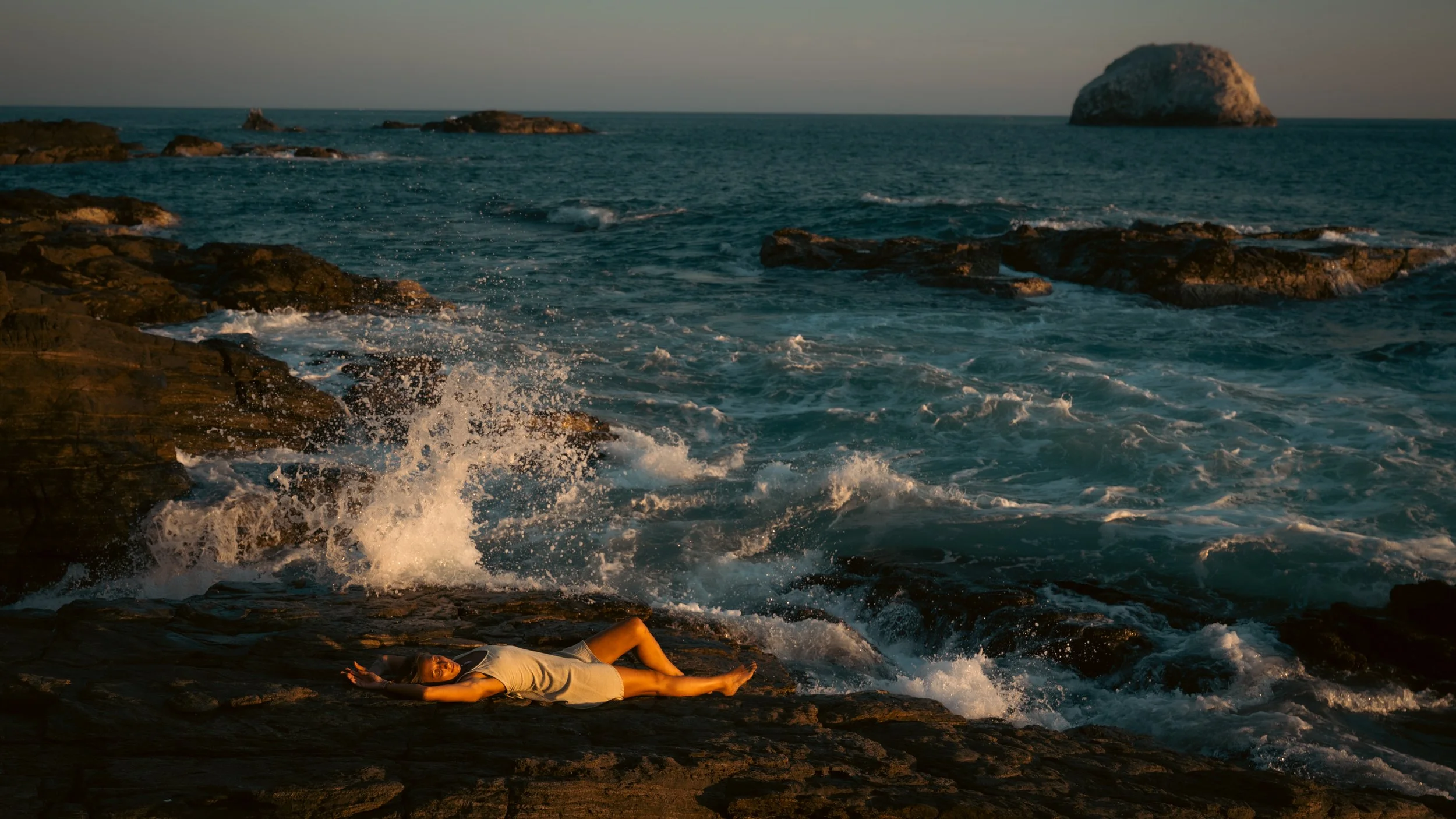 A woman lying on rocks near the ocean, with waves splashing around and large rocks and a distant rock formation in the background.