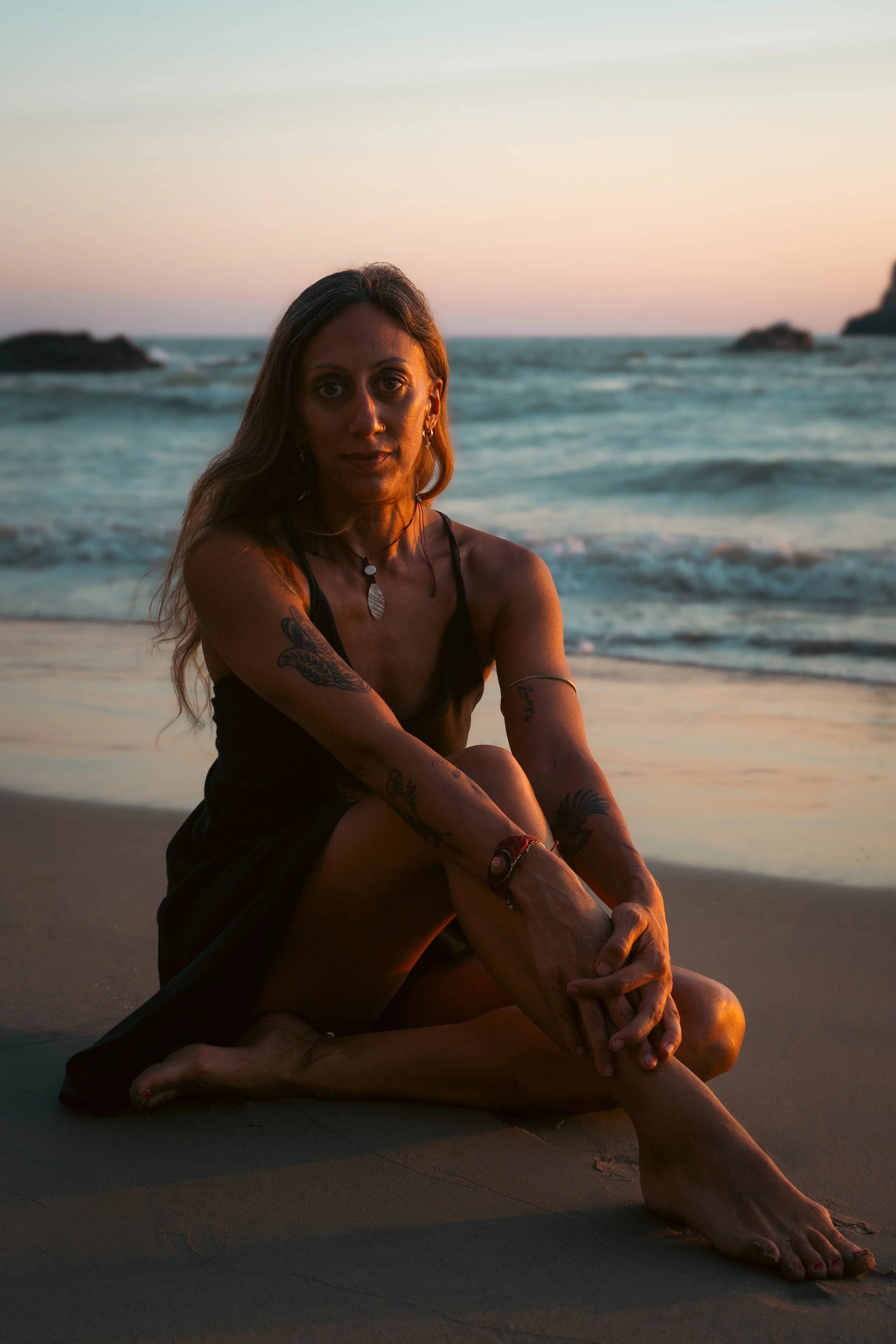 A woman with tattoos sits on the beach at sunset, with the ocean and rocks in the background.