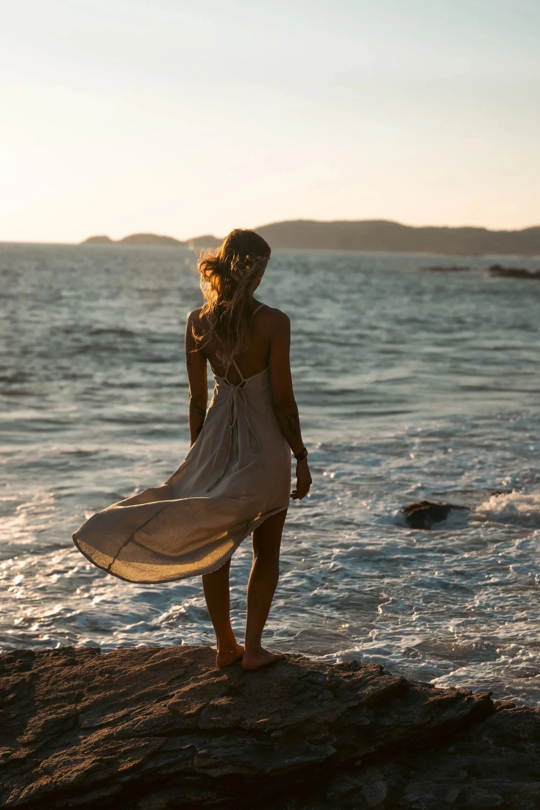 A woman wearing a light dress standing on a rock by the ocean at sunset, facing the water, with hills in the background.