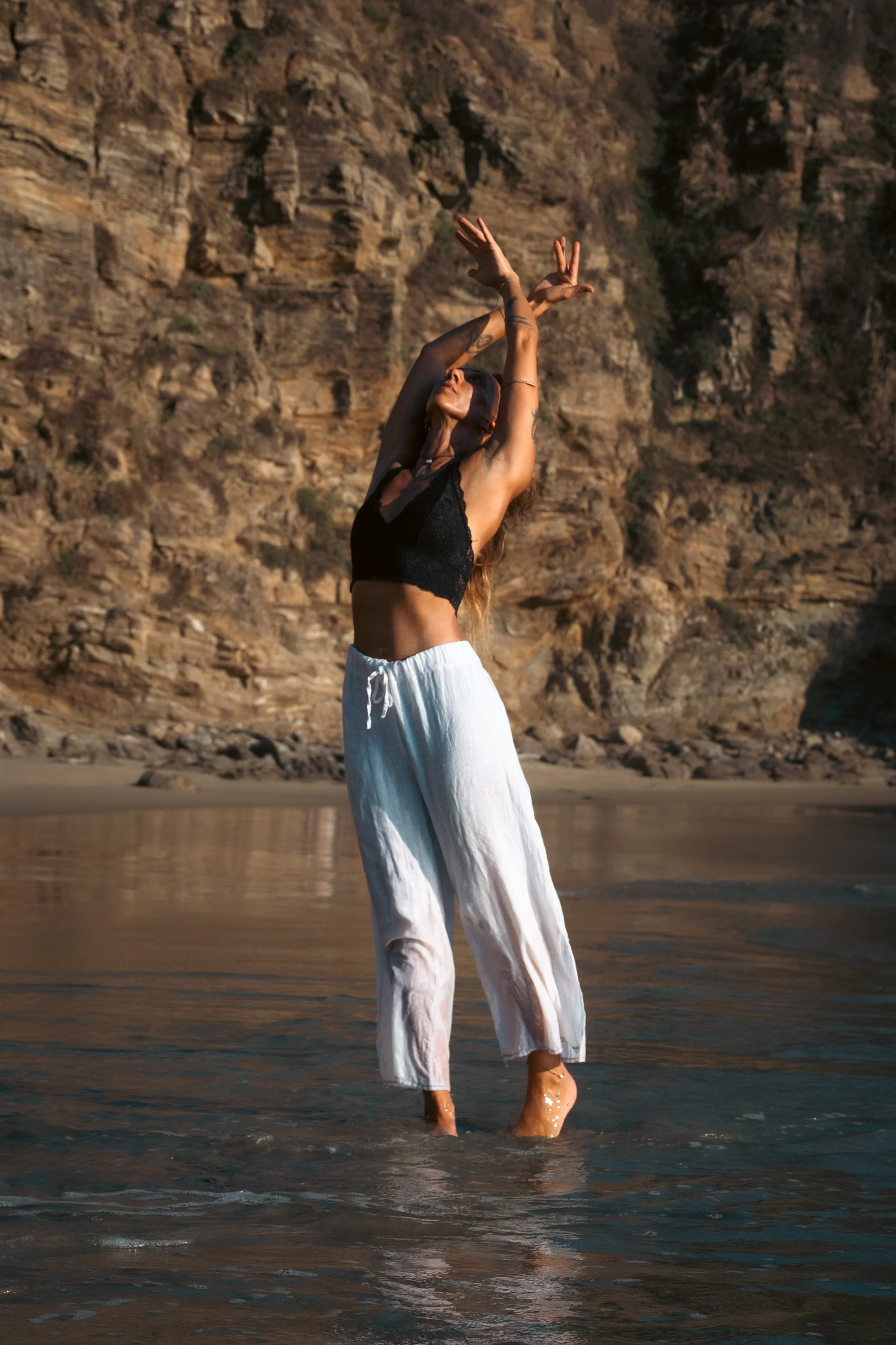 A woman standing in shallow water near a rocky cliff, wearing a black lace top and white pants, with arms raised and hair flowing.