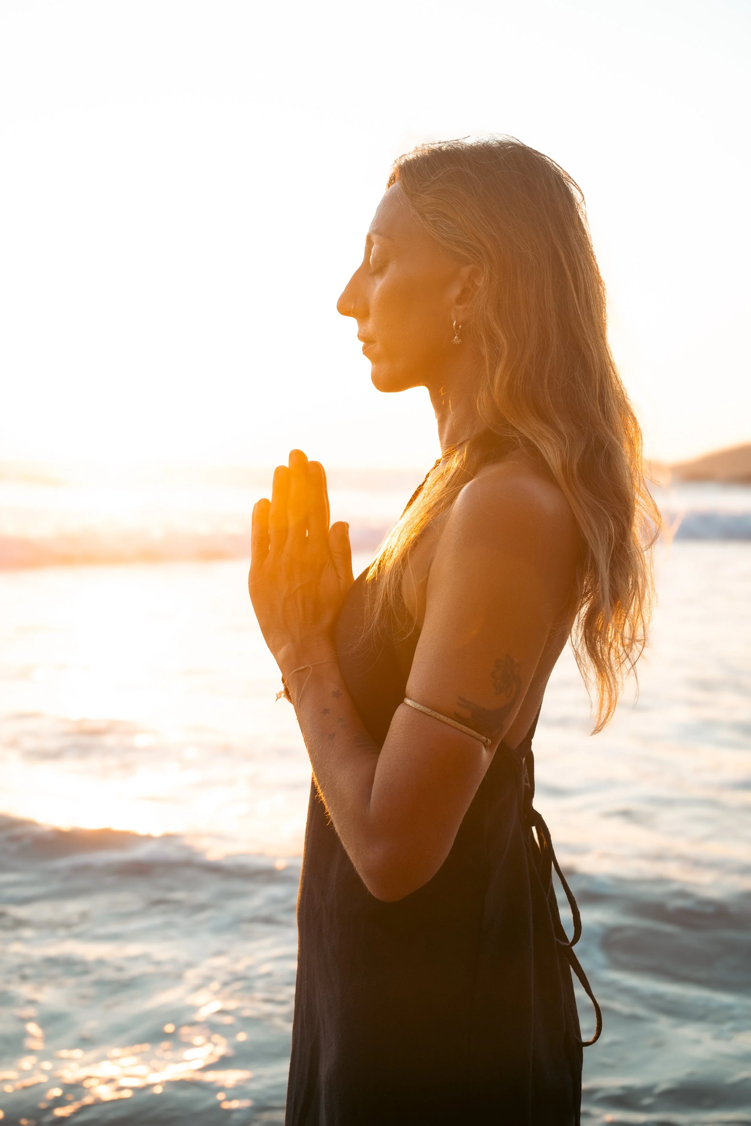 A woman with long, wavy hair practicing yoga on the beach at sunset, standing in profile with her hands pressed together in prayer position, eyes closed, facing the ocean.