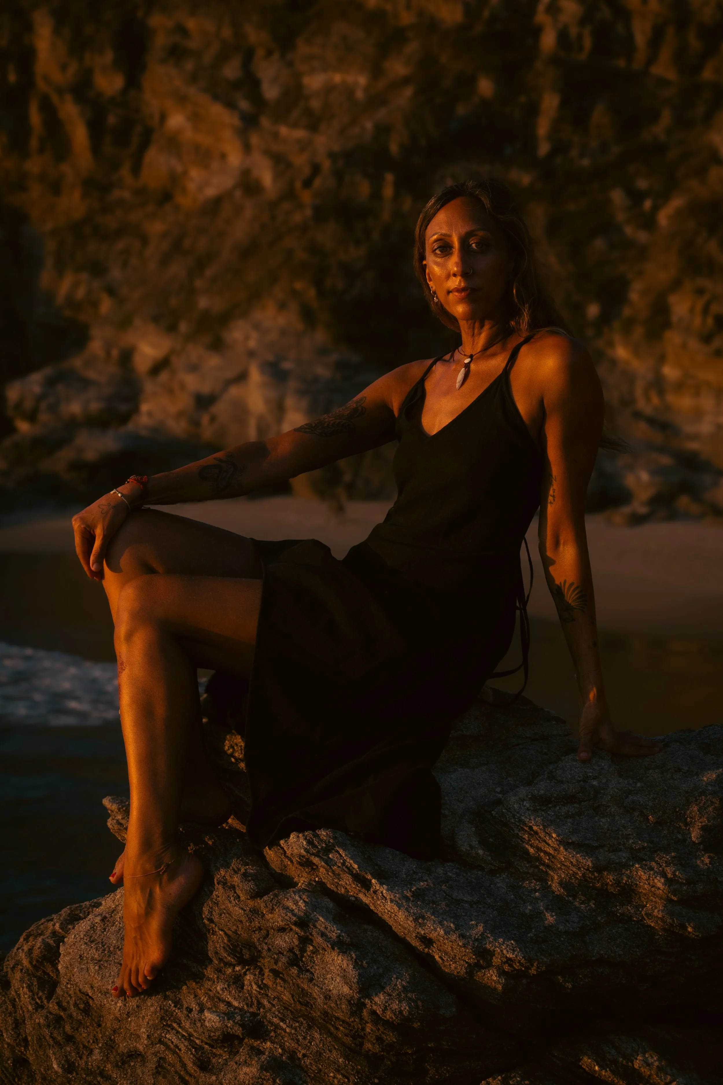 A woman sitting on a rock by the water during sunset, wearing a black dress and jewelry, with tattoos visible on her arm and leg.