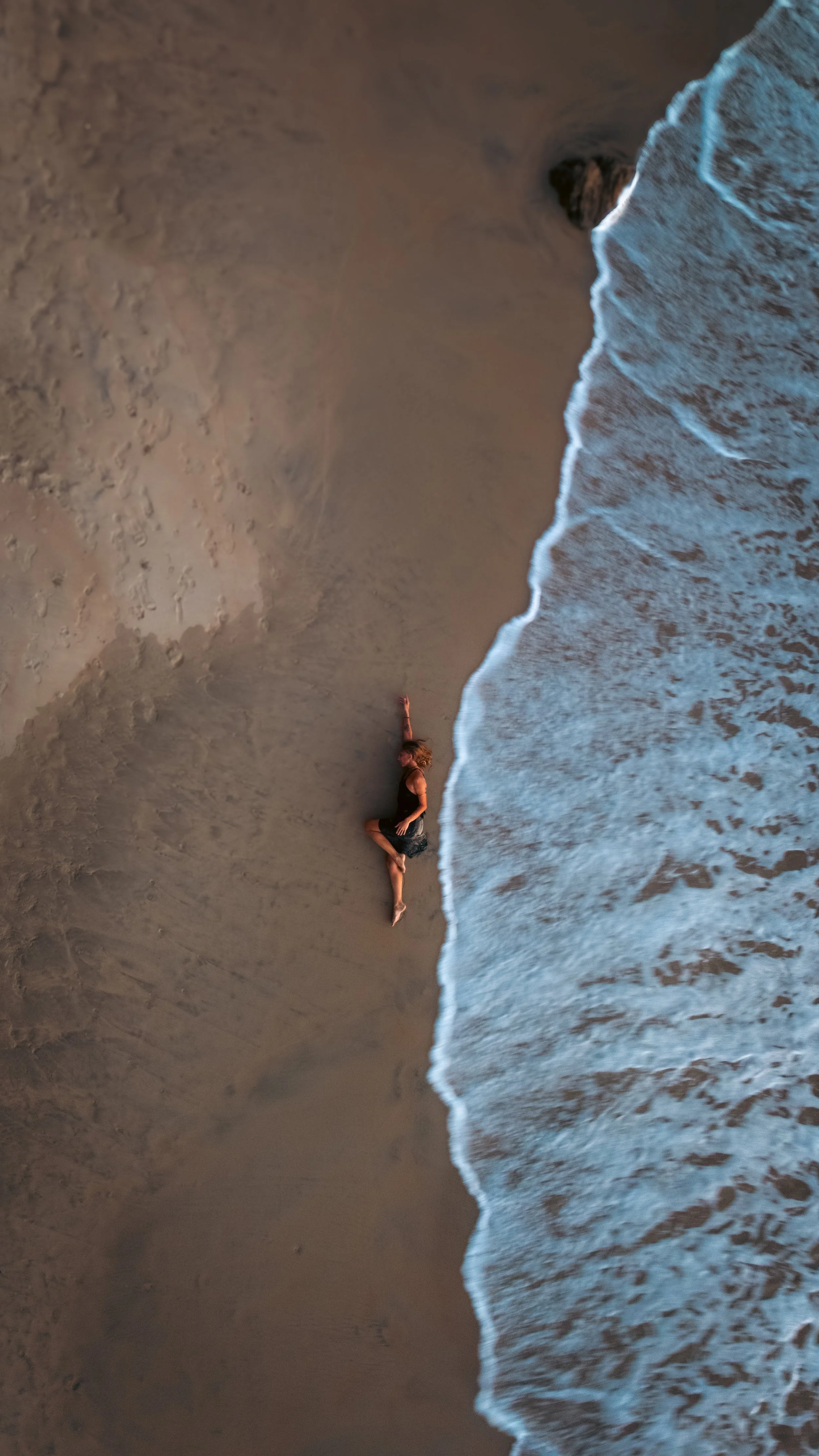 A person lying on the sandy beach near the water, with waves washing up close to them, seen from an aerial view.